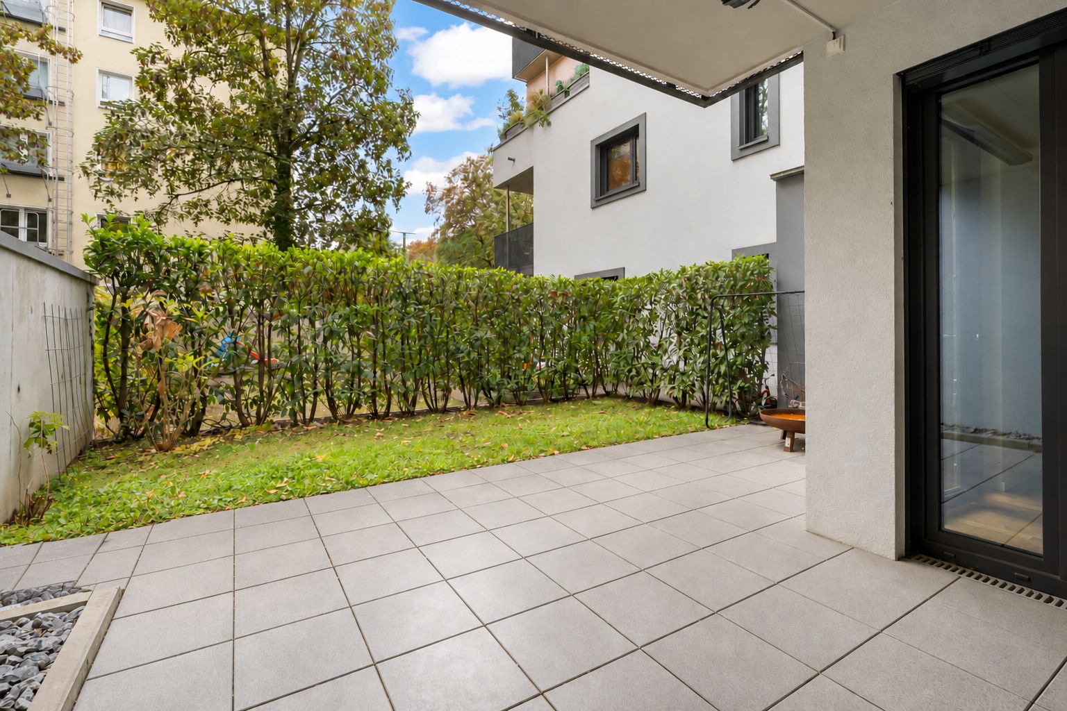 Überdachte Terrasse mit Fliesenboden und kleinem Garten, umgeben von einer Hecke.
