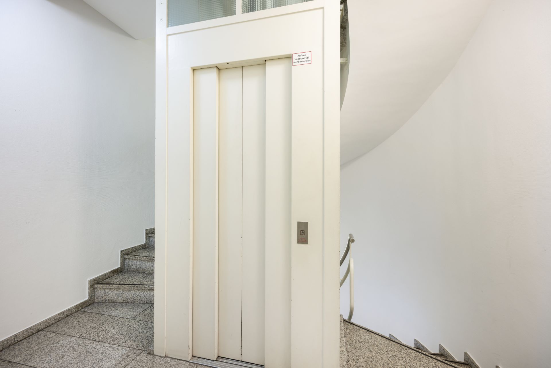 Geschlossener Aufzug mit weißen Türen neben einer gewendelten Steintreppe in einem Flur.