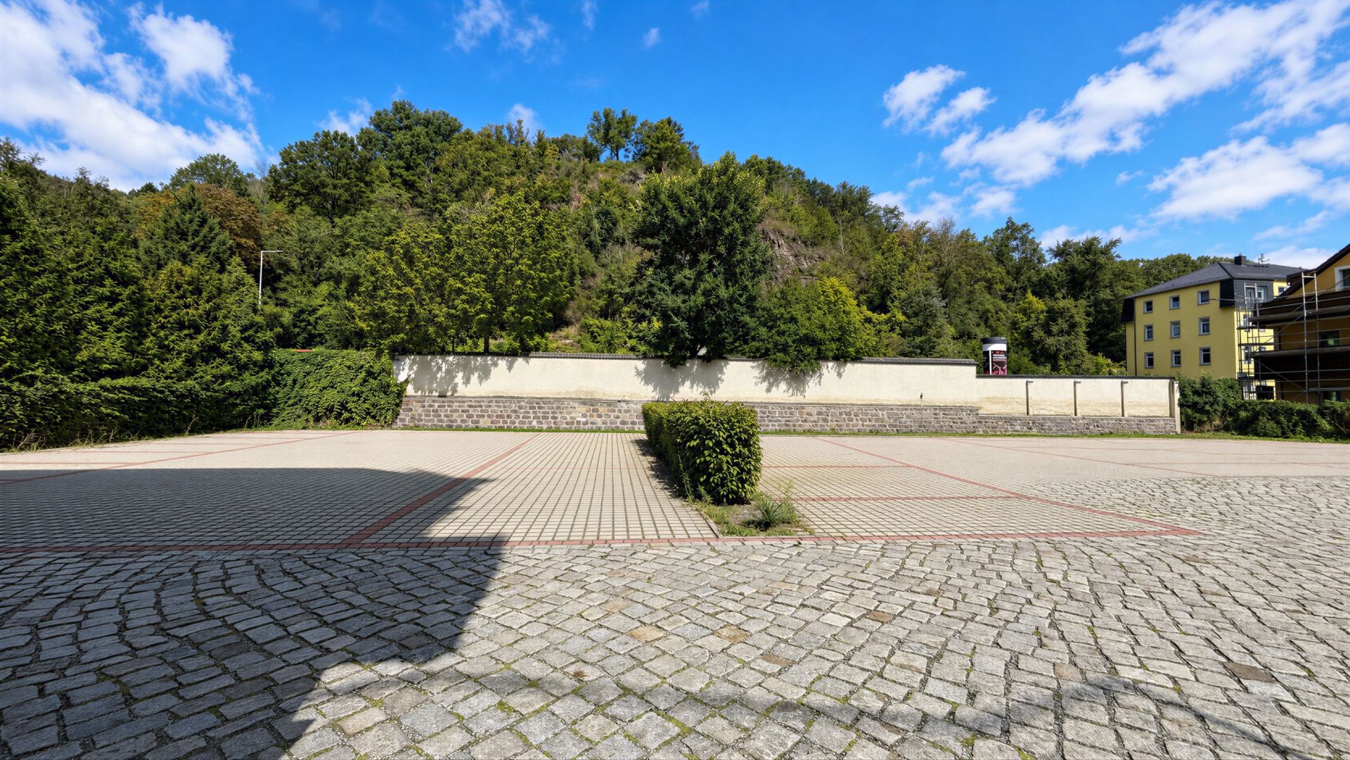 Gepflasterter Parkplatz mit einzelnen Büschen, im Hintergrund Bäume und ein gelbes Wohnhaus.