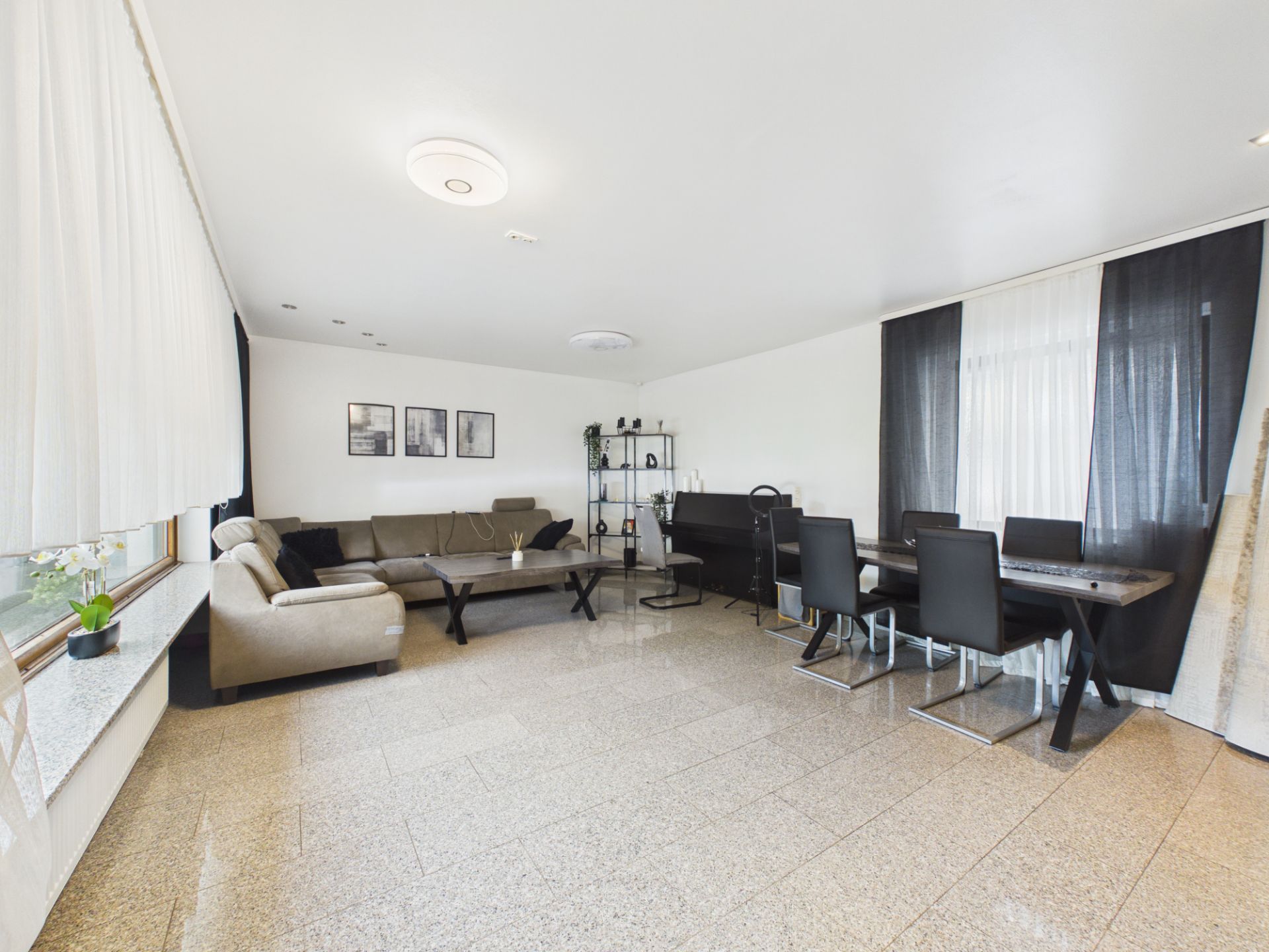 Helles Wohnzimmer mit Ecksofa, Esstisch, Stühlen und großen Fenstern mit Vorhängen.
