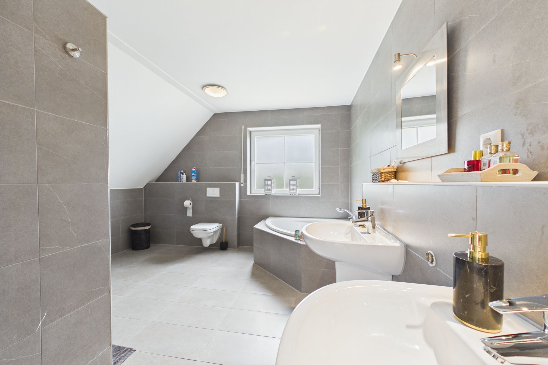 Modernes Badezimmer mit Badewanne, zwei Waschbecken, WC und grauen Fliesen, Fenster mittig an der Wand.