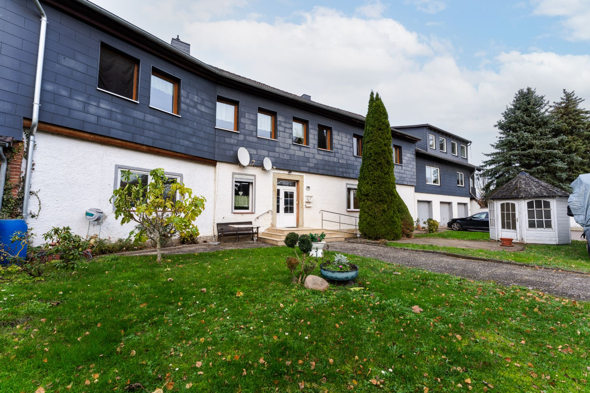 Zweistöckiges Wohnhaus mit Garten, Sat-Schüsseln und kleinem Pavillon im Vorgarten.