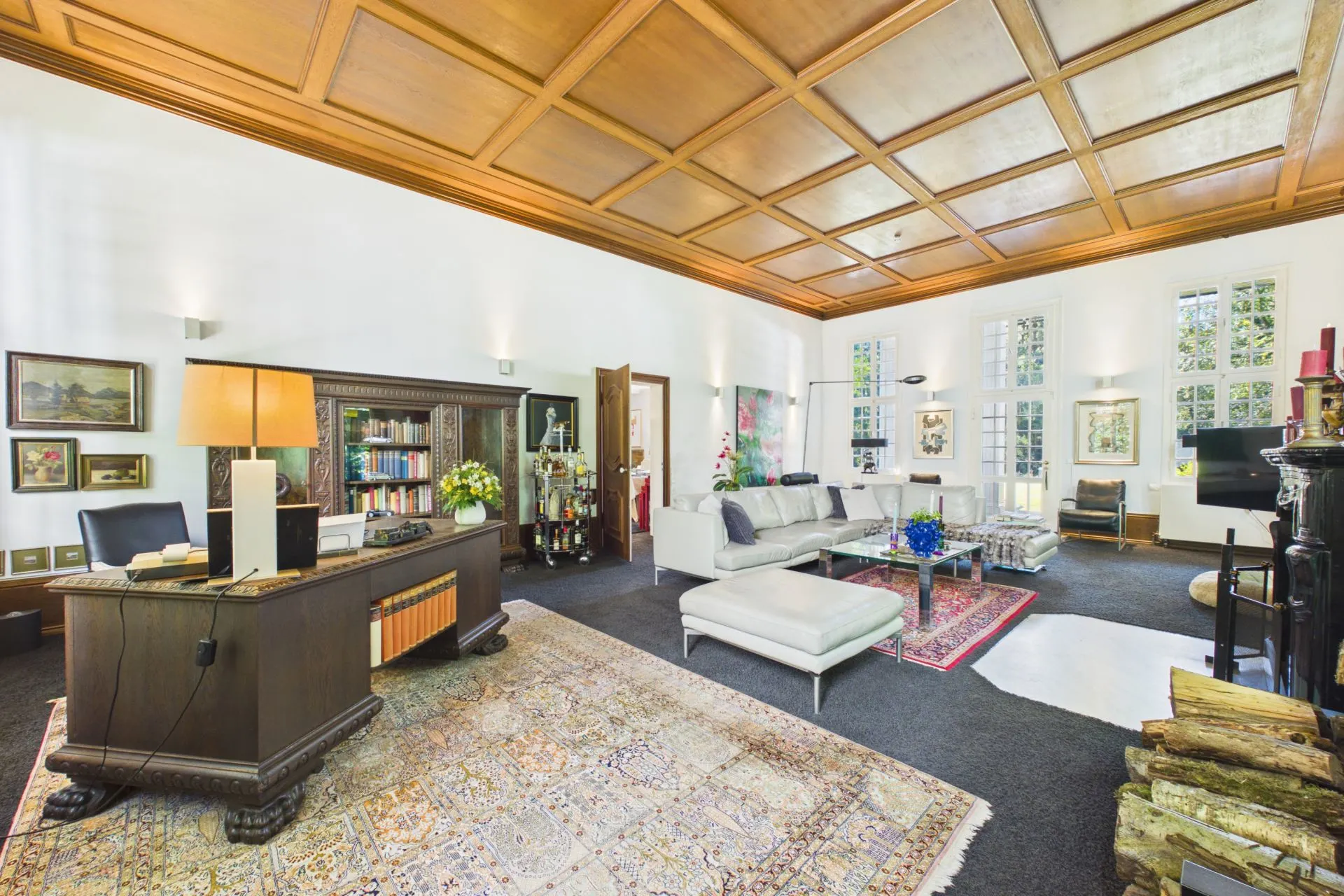 Großes Wohnzimmer mit Holzdecke, Schreibtisch, Bücherregal, Sofas und großen Fenstern.
