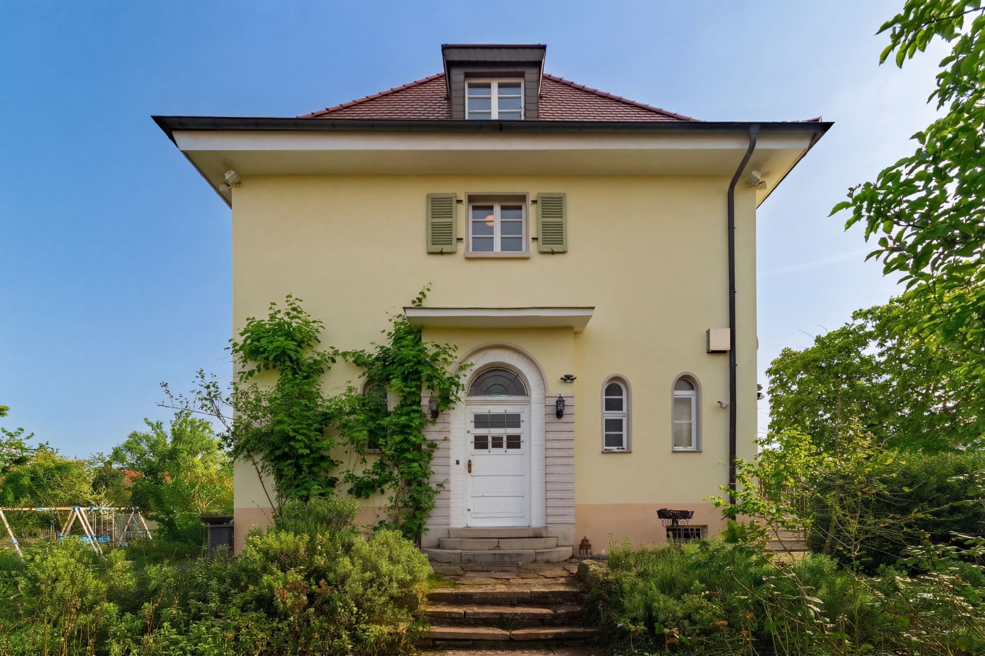Gelbes Wohnhaus mit rotem Dach, zentraler Haustür und begrüntem Eingangsbereich.