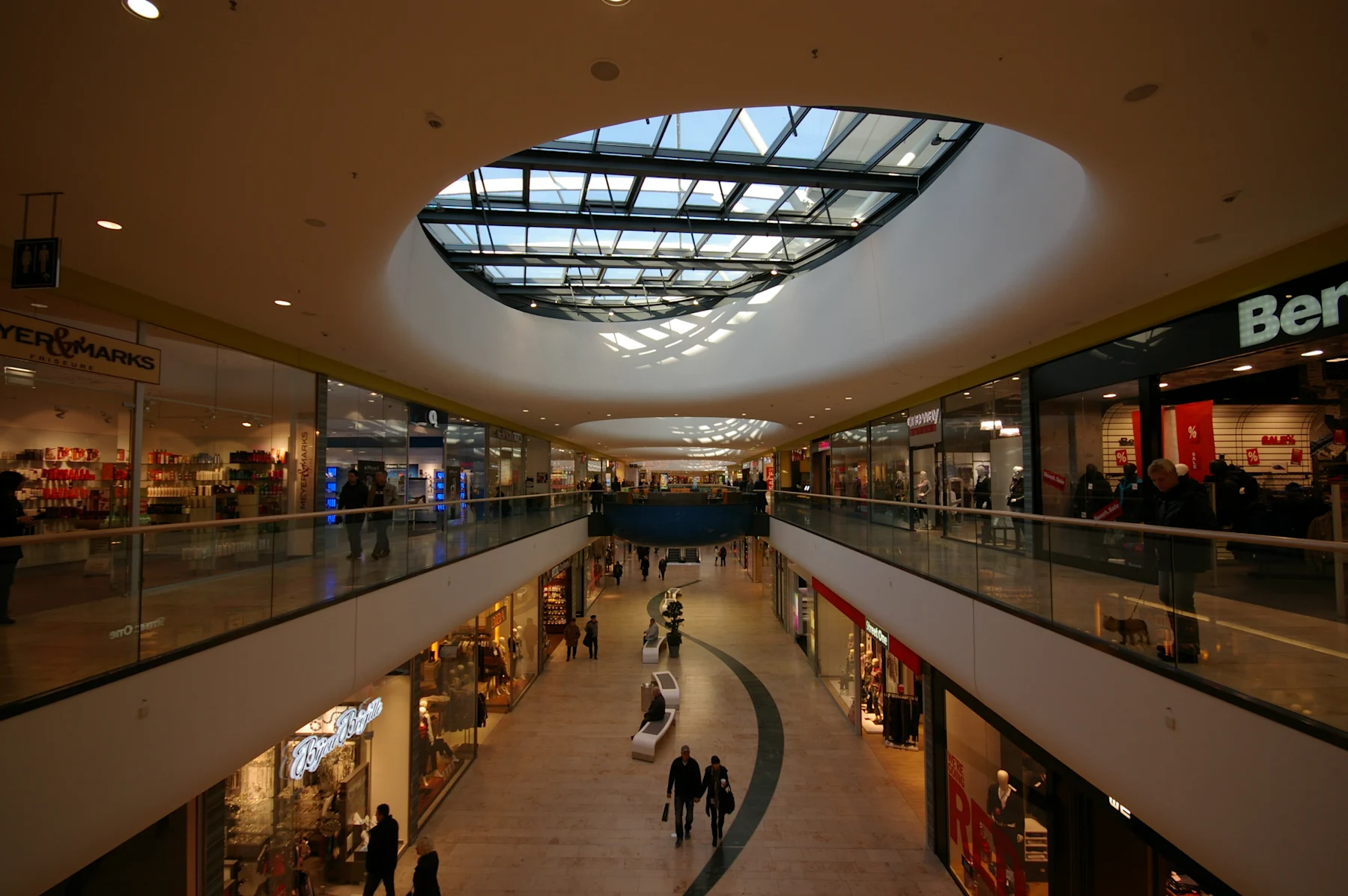 Innenansicht eines Einkaufszentrums mit Glasdach und Läden auf zwei Etagen.