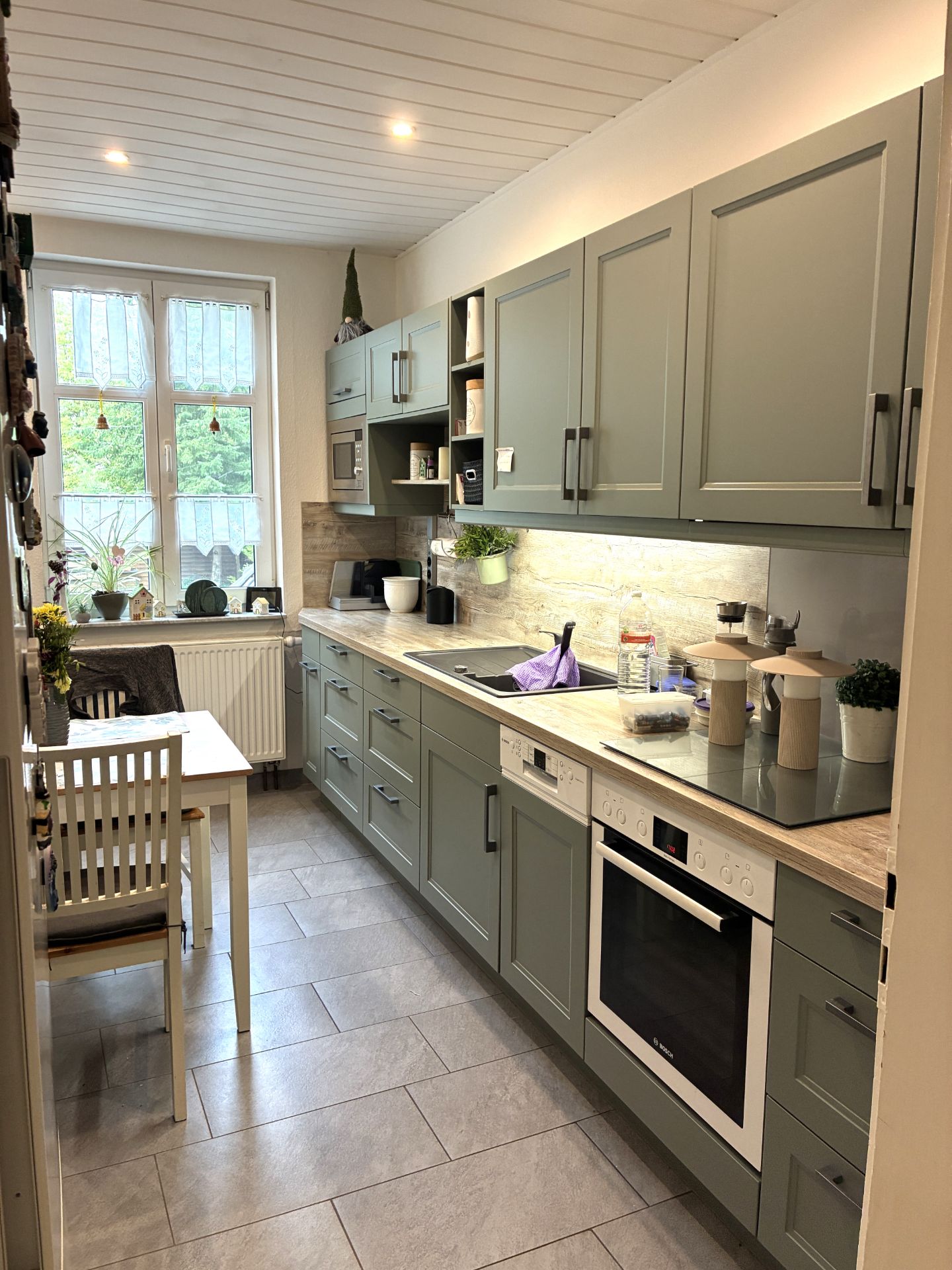 Moderne Küche mit grünen Schränken, Esstisch, großem Fenster und Elektrogeräten.
