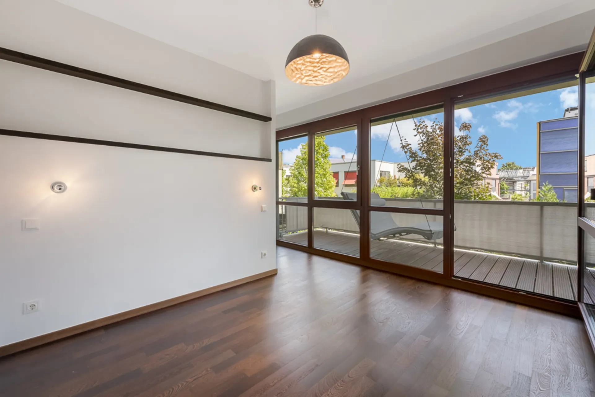 Leeres Zimmer mit Holzboden, großen Fenstern und Zugang zu einem Balkon.