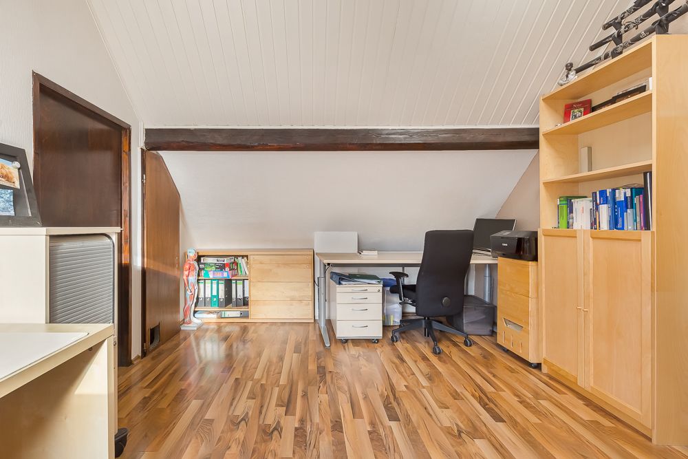 Helles Arbeitszimmer mit Holzschrank, Schreibtisch, Stuhl und Regalen unter Dachschräge.