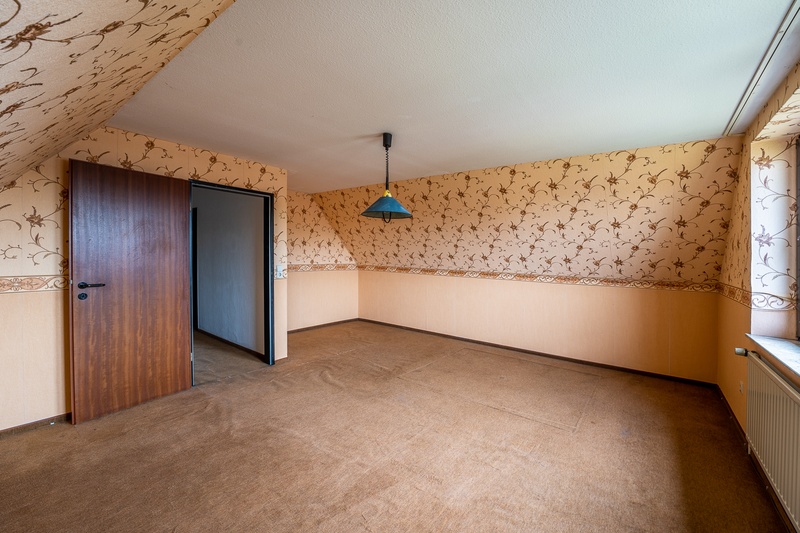 Leeres Zimmer mit gemusterter Tapete, braunem Teppichboden und offenstehender Holztür.