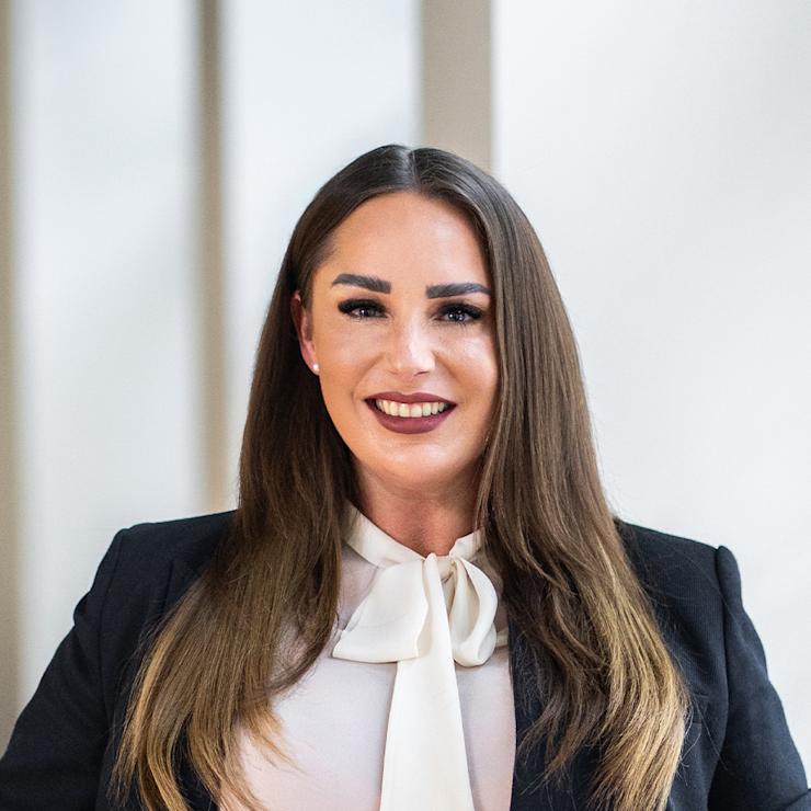 Frau in weißer Bluse mit Schleife und schwarzem Blazer steht lächelnd vor hellem Hintergrund.