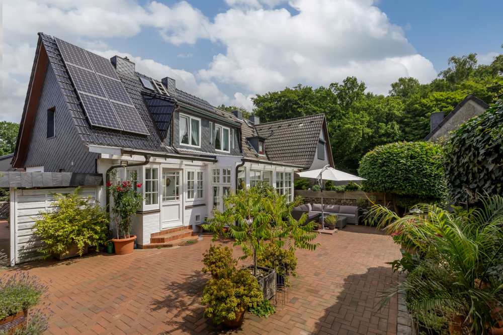 Haus mit Solarpanelen, großer Terrasse, vielen Pflanzen und Gartenmöbeln unter Sonnenschirm.