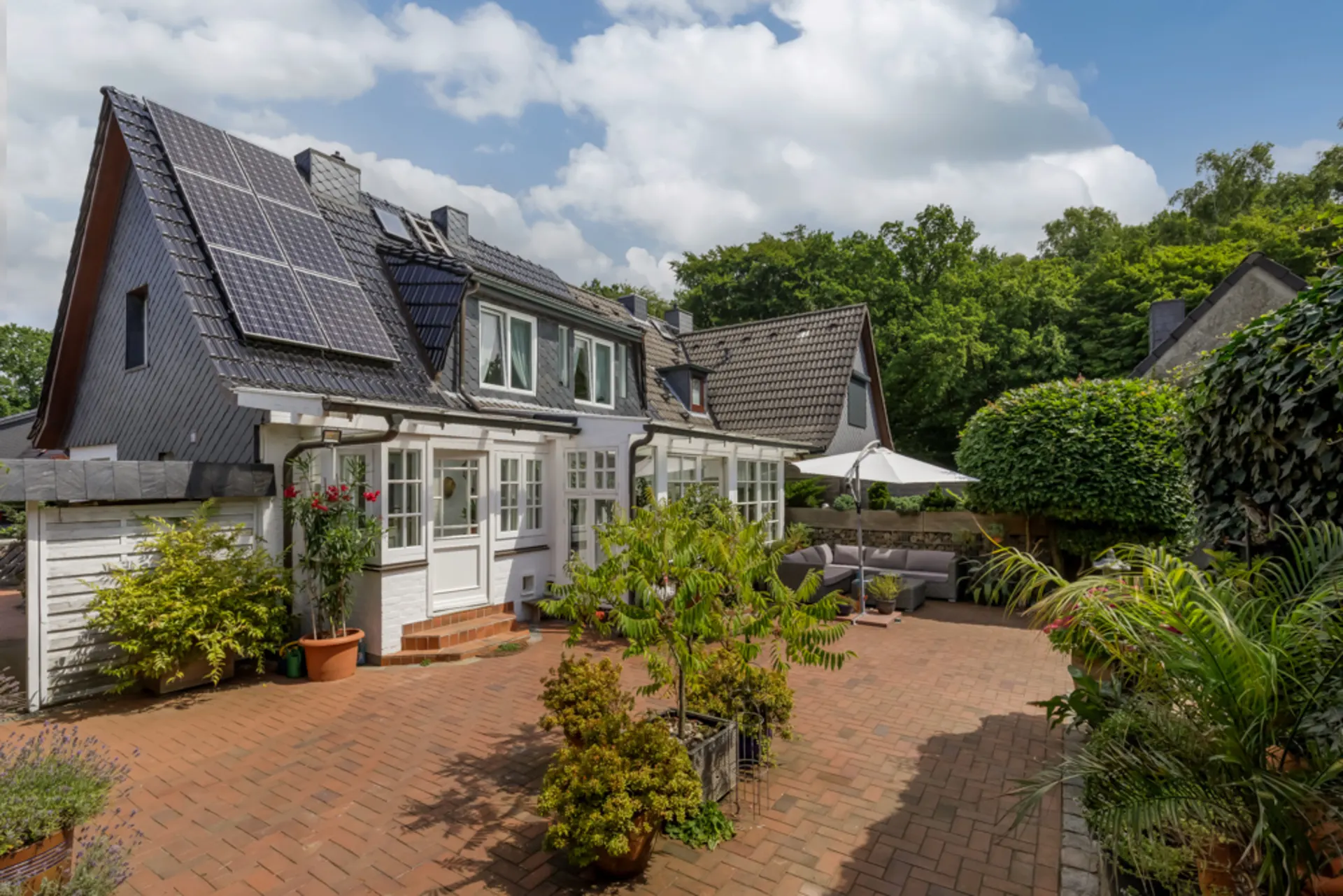 Haus mit Solarpanelen, großer Terrasse, vielen Pflanzen und Gartenmöbeln unter Sonnenschirm.