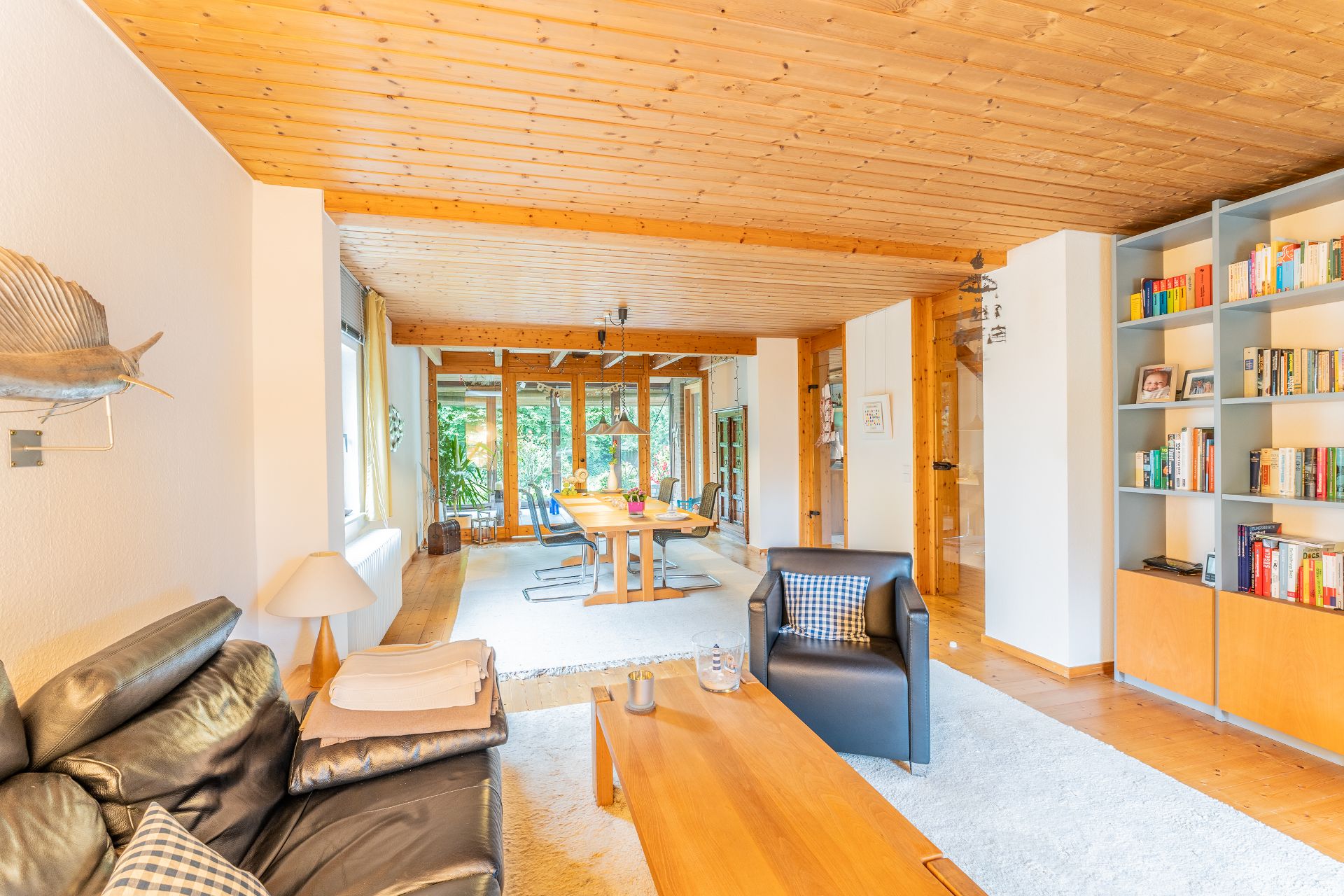 Helles Wohnzimmer mit Holzboden, Ledersofa, Esstisch, Bücherregal und großen Fenstern zum Garten.