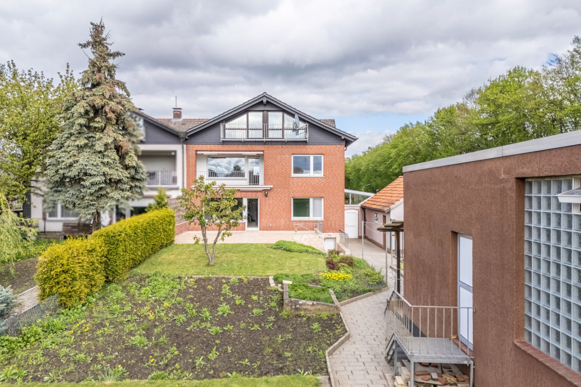 Zweistöckiges Backsteinhaus mit Garten, Bäumen und Nebengebäude im Vordergrund.