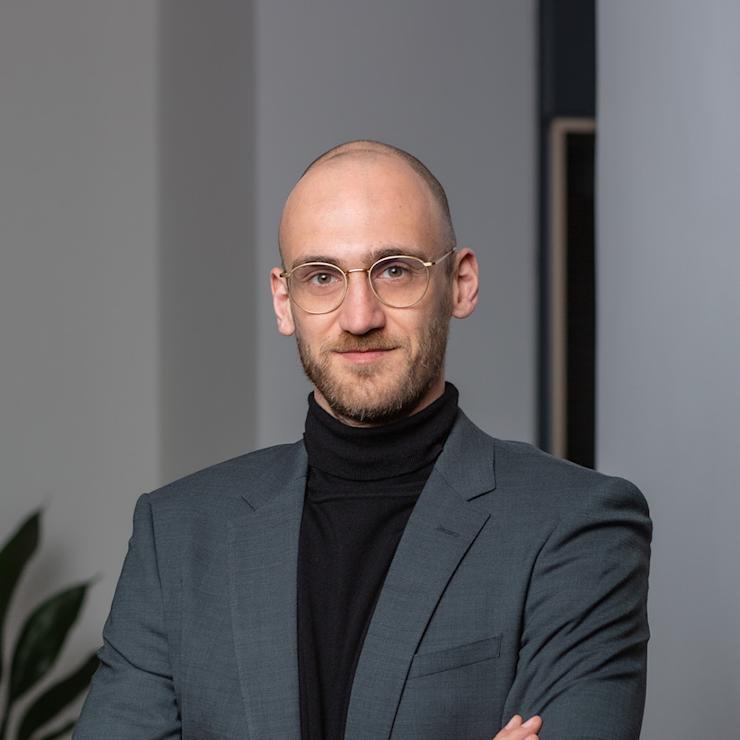 Ein Mann mit Brille und Bart trägt einen Anzug und steht mit verschränkten Armen vor einer Pflanze.