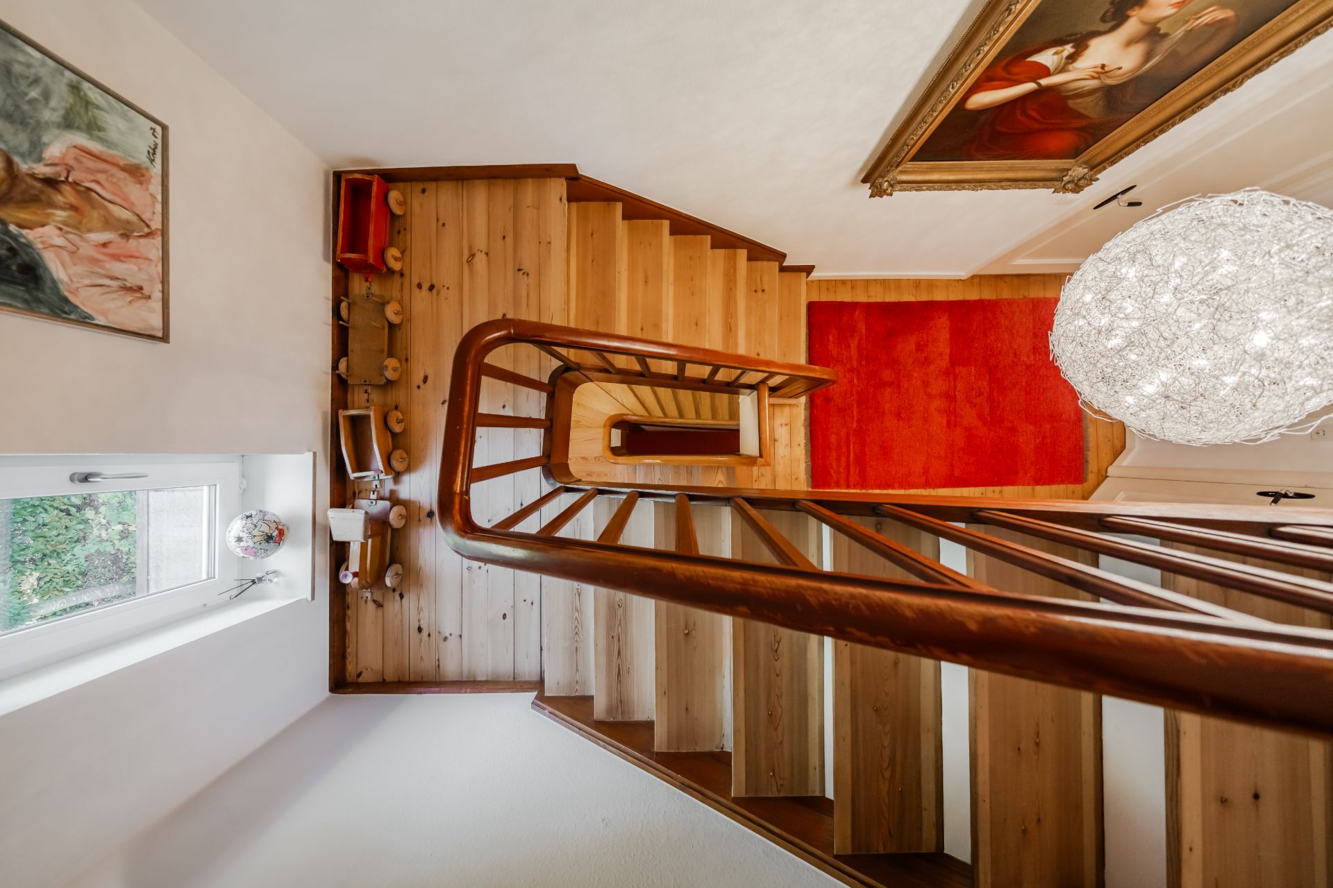 Holztreppe mit Handlauf, rotem Teppich, Gemälden an der Wand und Fenster mit Blick nach draußen.