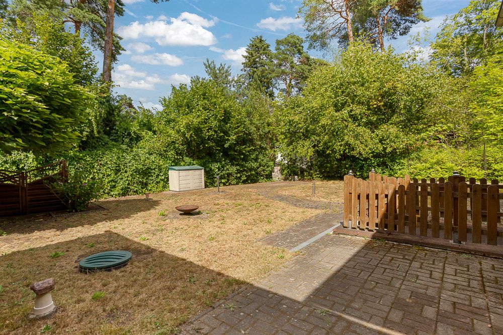 Garten mit Rasenfläche, einigen Gegenständen, Terrasse und Holzzaun, umgeben von Bäumen.