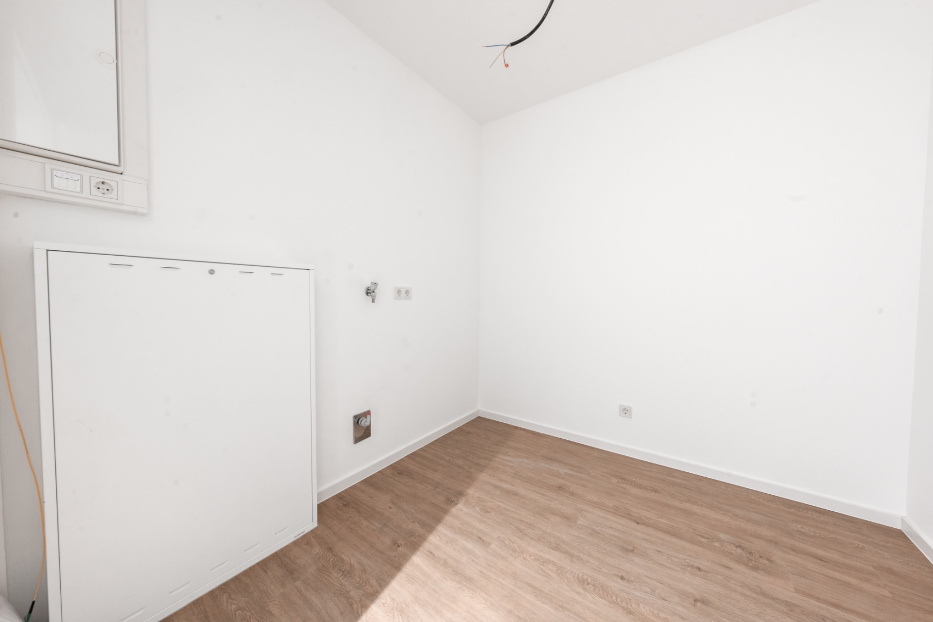 Leerer, heller Raum mit weißem Wänden und hellem Holzboden, Wandanschlüssen und Kabel von der Decke.