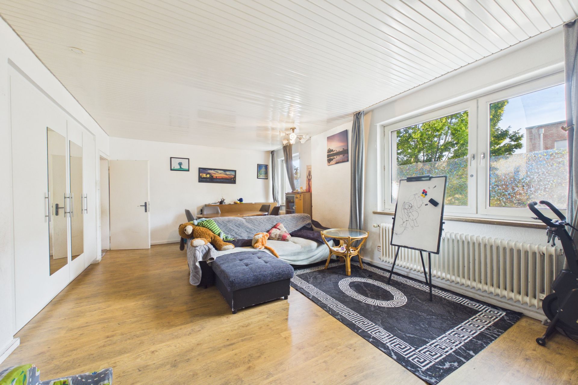 Helles Wohnzimmer mit großem Sofa, Teppich, Whiteboard und Fenster mit Blick nach draußen.