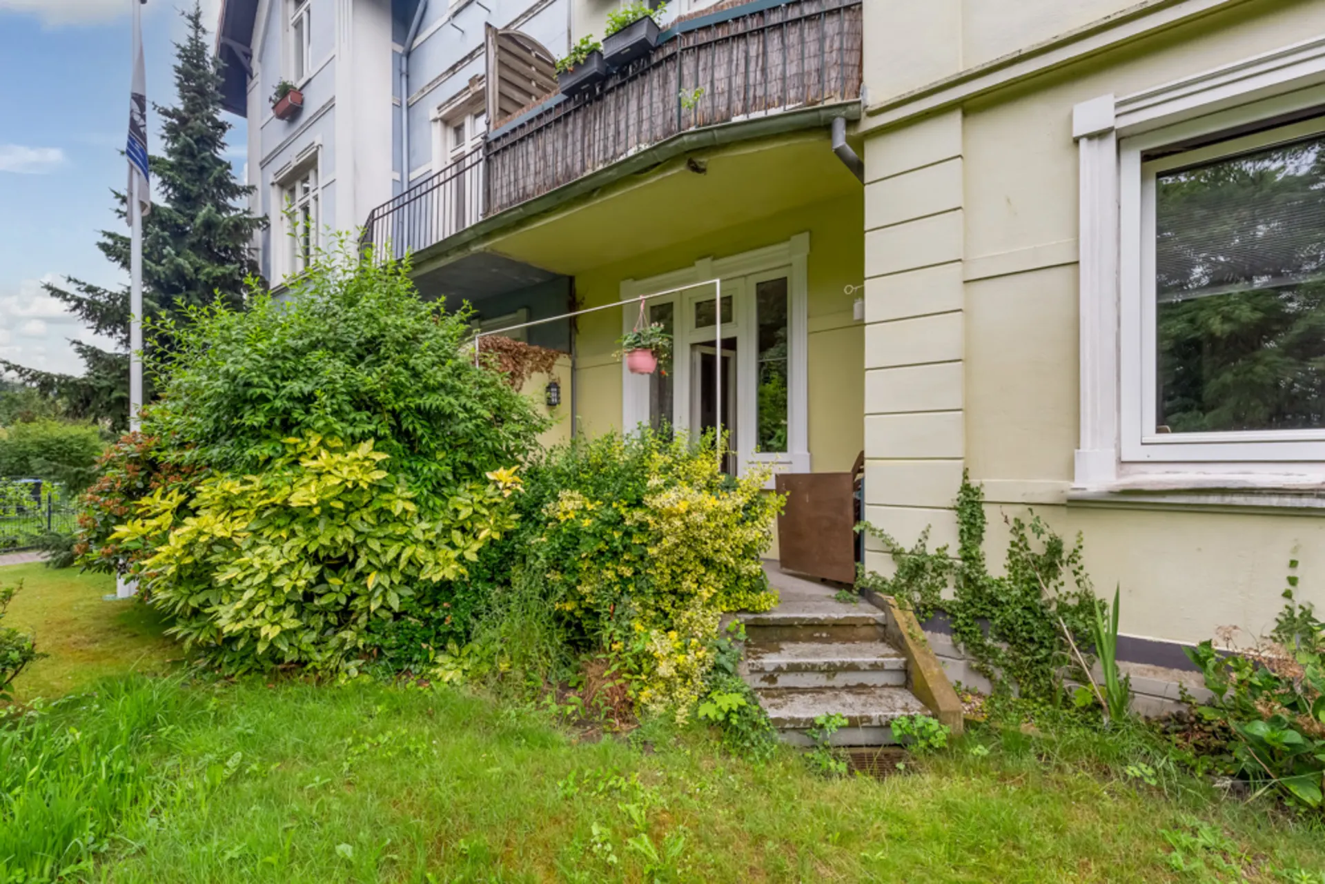 Eingang mit kleiner Treppe, umgeben von Sträuchern und grüner Wiese vor einem Wohnhaus.