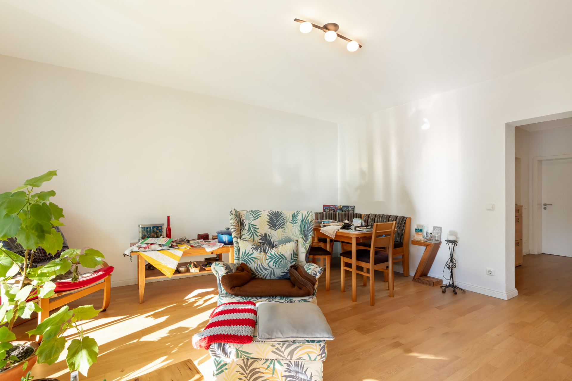 Helles Wohnzimmer mit Sessel, Esstisch, Stühlen, Pflanzen und Holzboden.