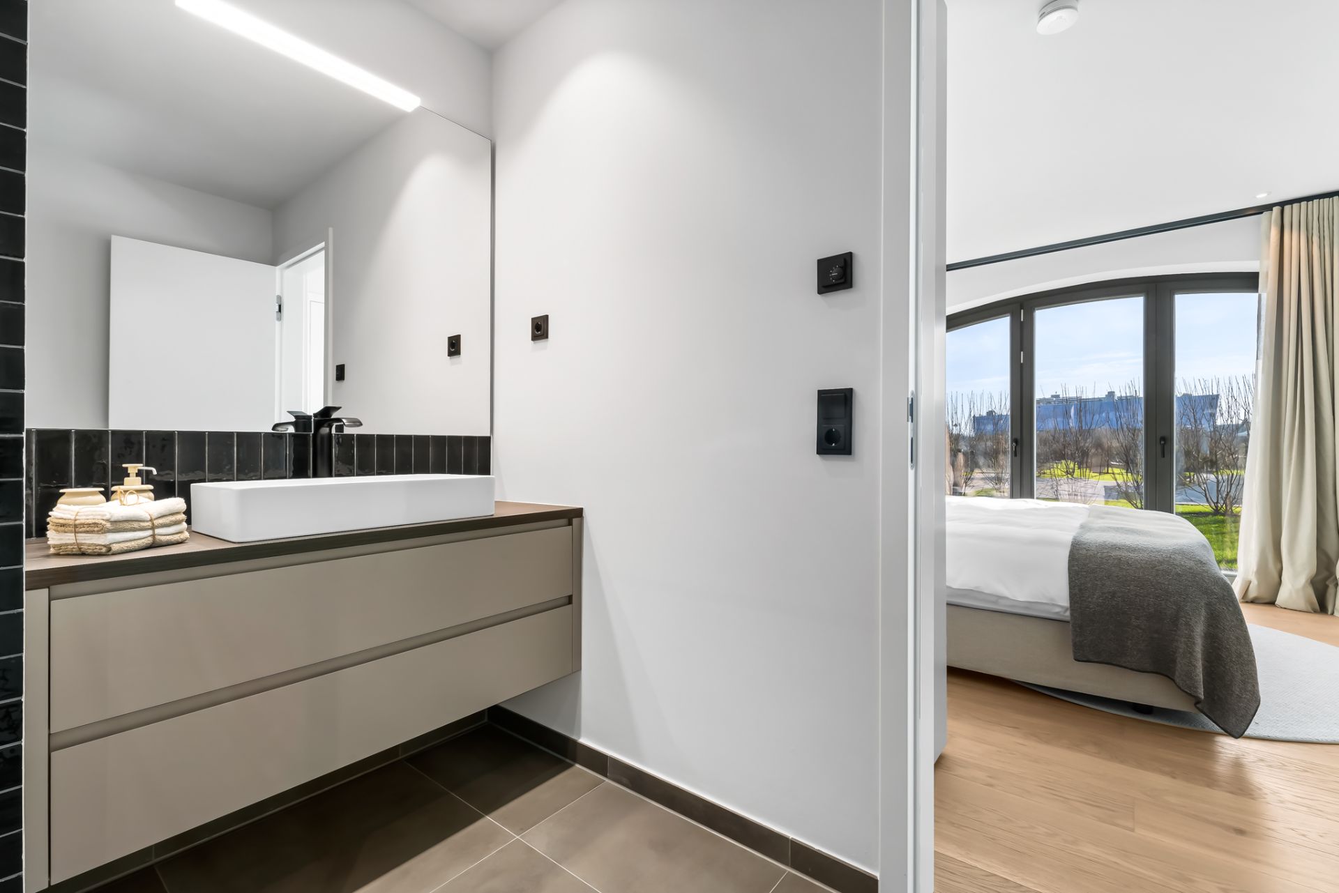 Modernes Badezimmer mit großem Spiegel und Waschbecken, angrenzend an ein helles Schlafzimmer mit Fenster.