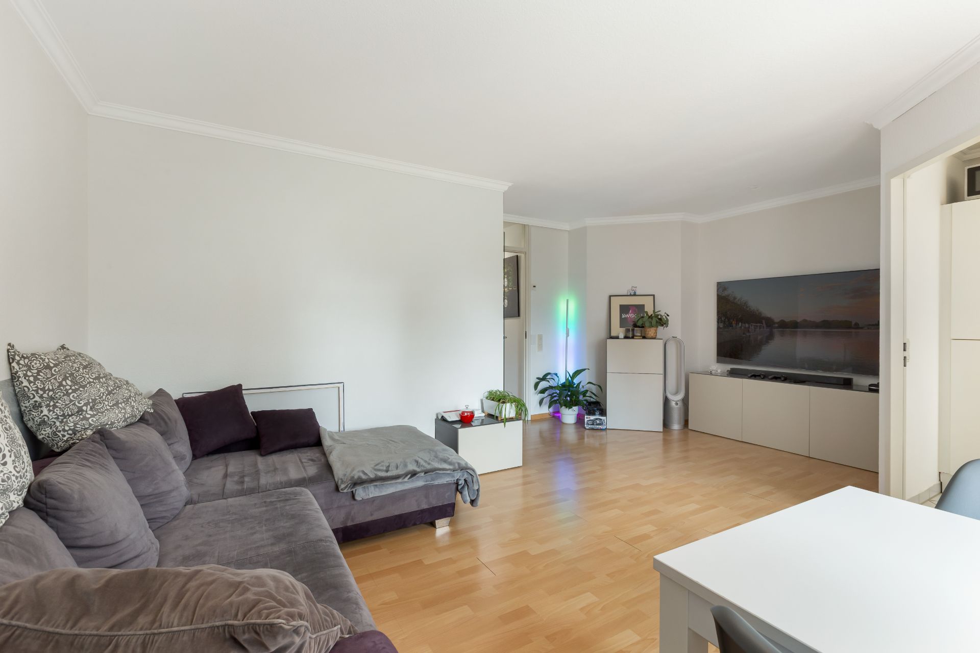Helles Wohnzimmer mit grauem Sofa, Holzboden, kleinem Tisch und Fernseher an der Wand.