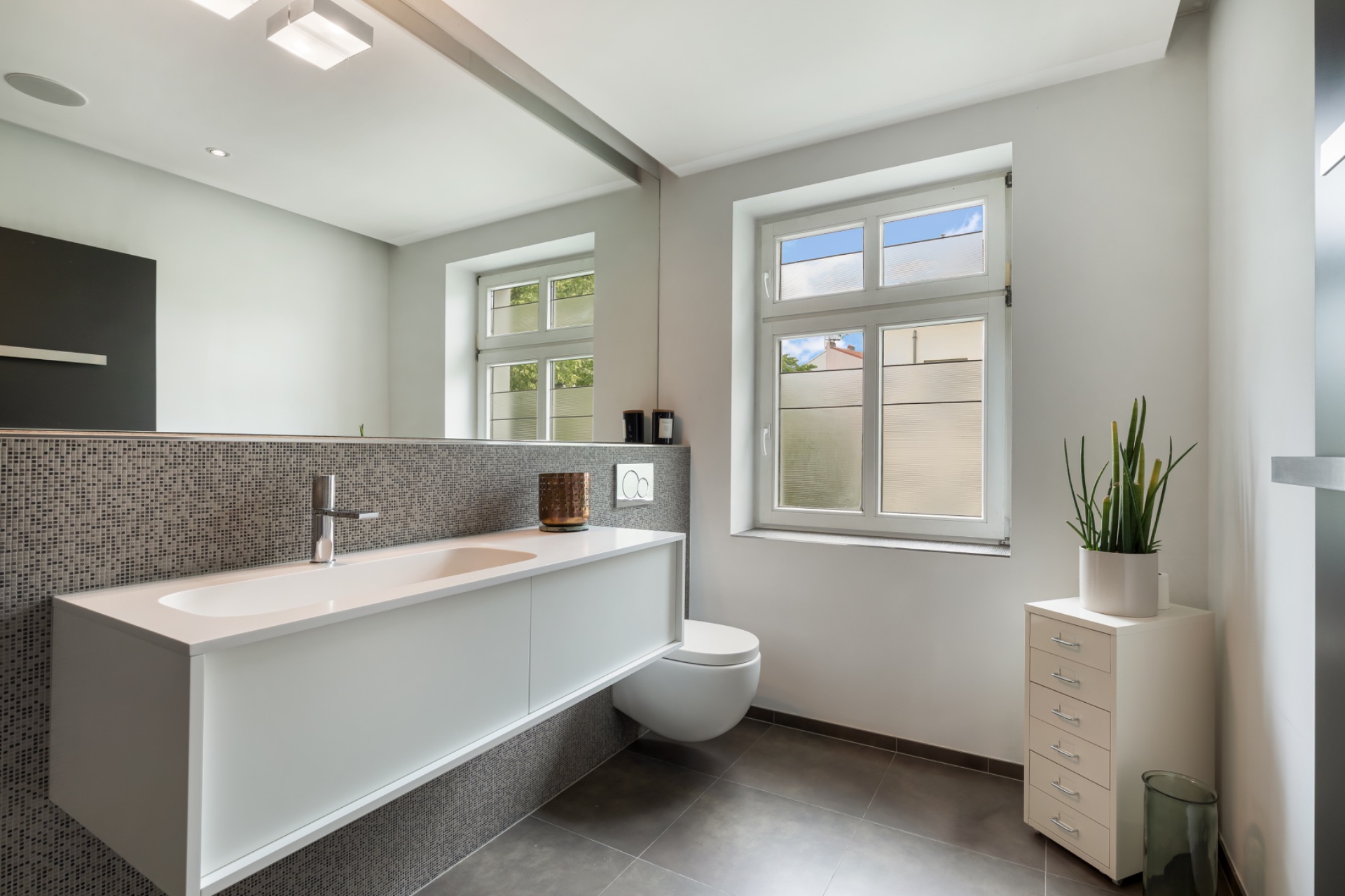 Modernes Badezimmer mit großem Spiegel, Waschbecken, Hänge-WC und Pflanze am Fenster.