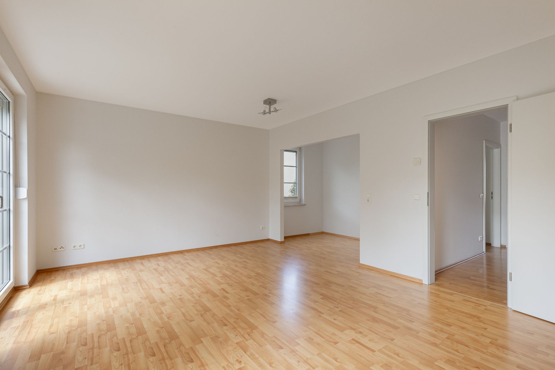 Heller leerer Raum mit Holzfußboden, weißen Wänden, Fenstern und offener Tür.