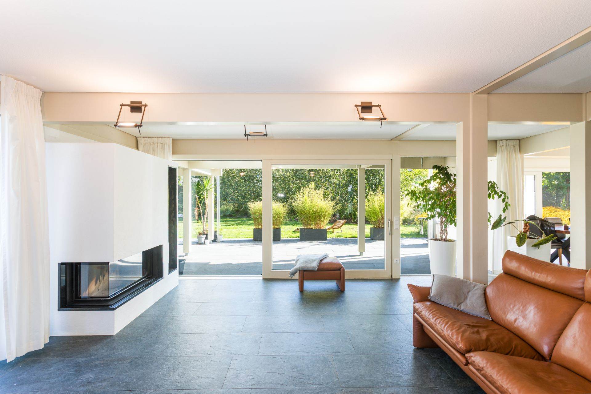 Helles Wohnzimmer mit braunem Ledersofa, Kamin und großen Fenstern zum Garten.