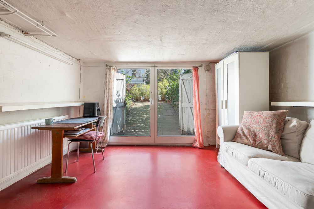 Heller Raum mit rotem Boden, Sofa, Tisch, Stühlen, Schrank und Glastüren zum Garten.