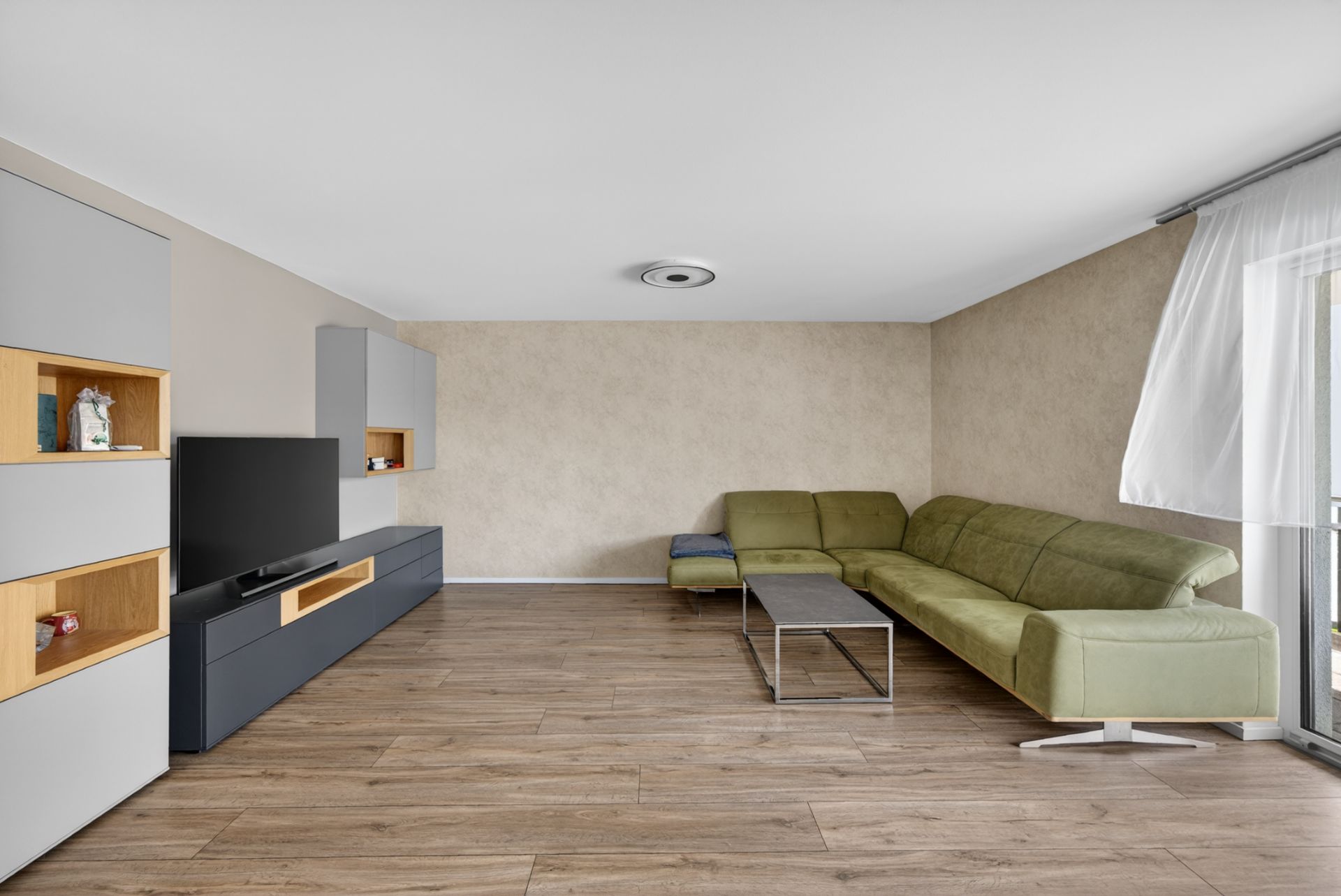 Wohnzimmer mit grüner Eckcouch, Couchtisch, Fernseher und Regal auf Holzfußboden.