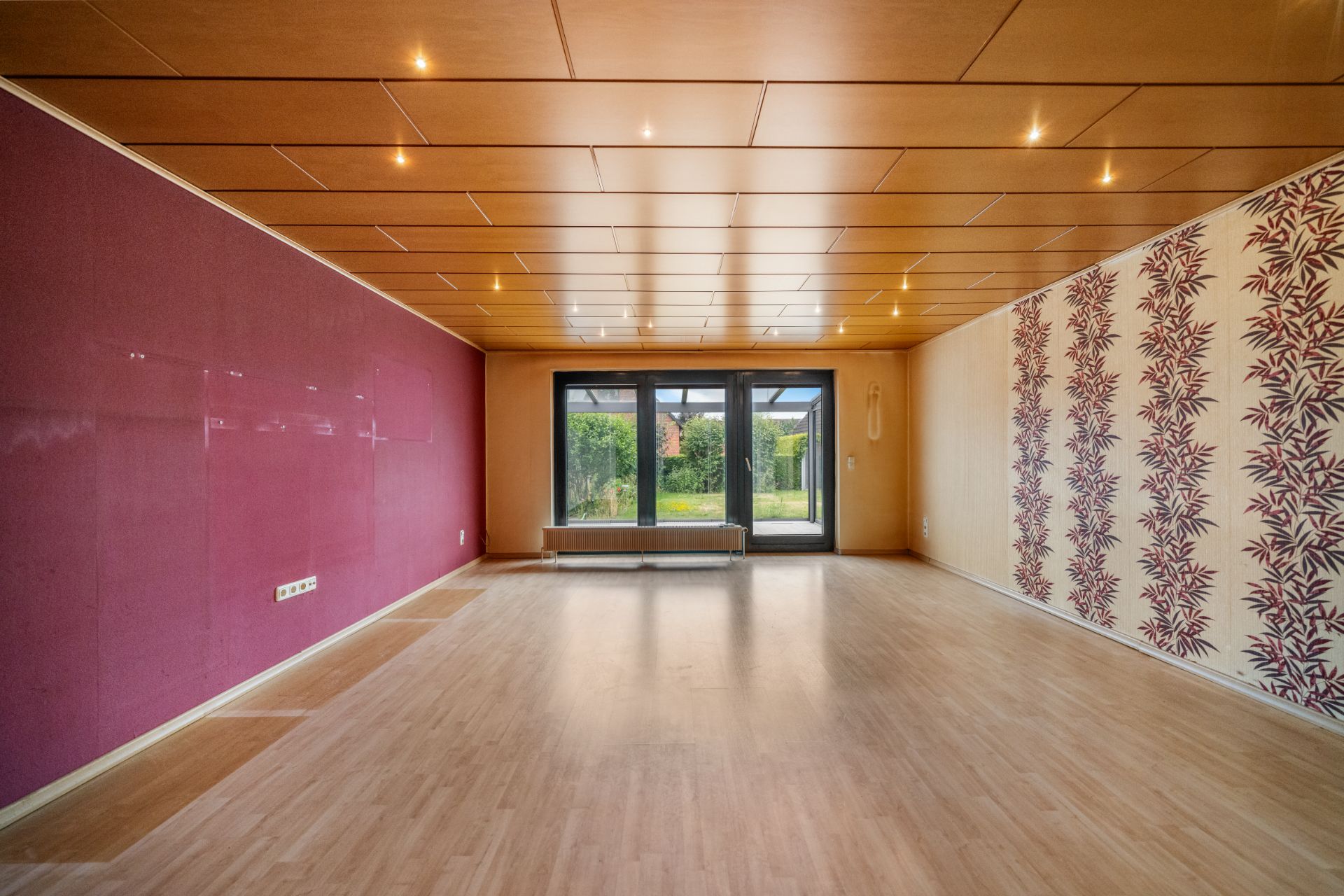 Leerstehendes Wohnzimmer mit Holzboden, bunter Tapete und großen Fenstern zum Garten.