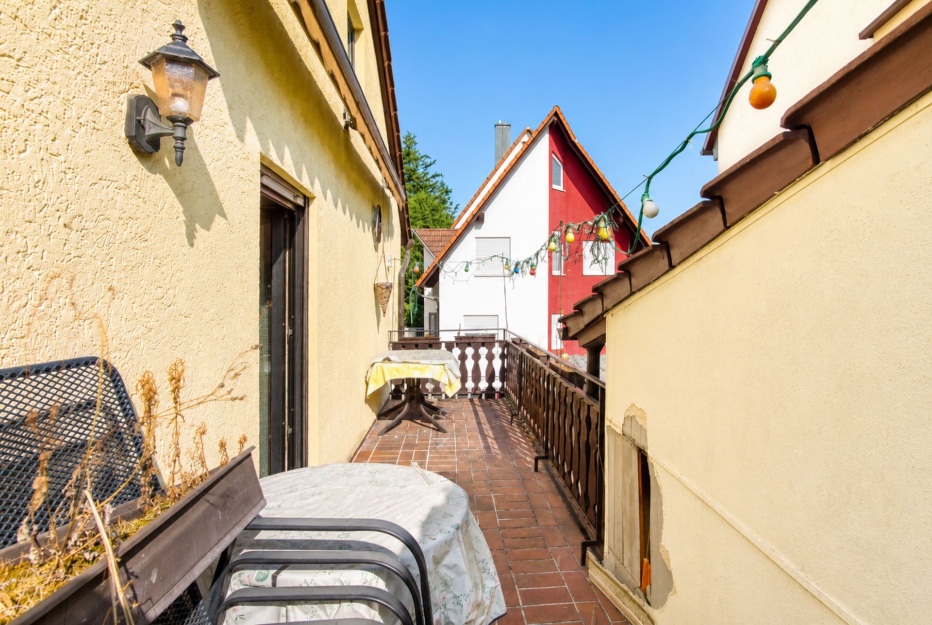 Sonnige Terrasse mit Tisch, Stühlen, Balustrade und Lichterkette zwischen gelben Hauswänden.