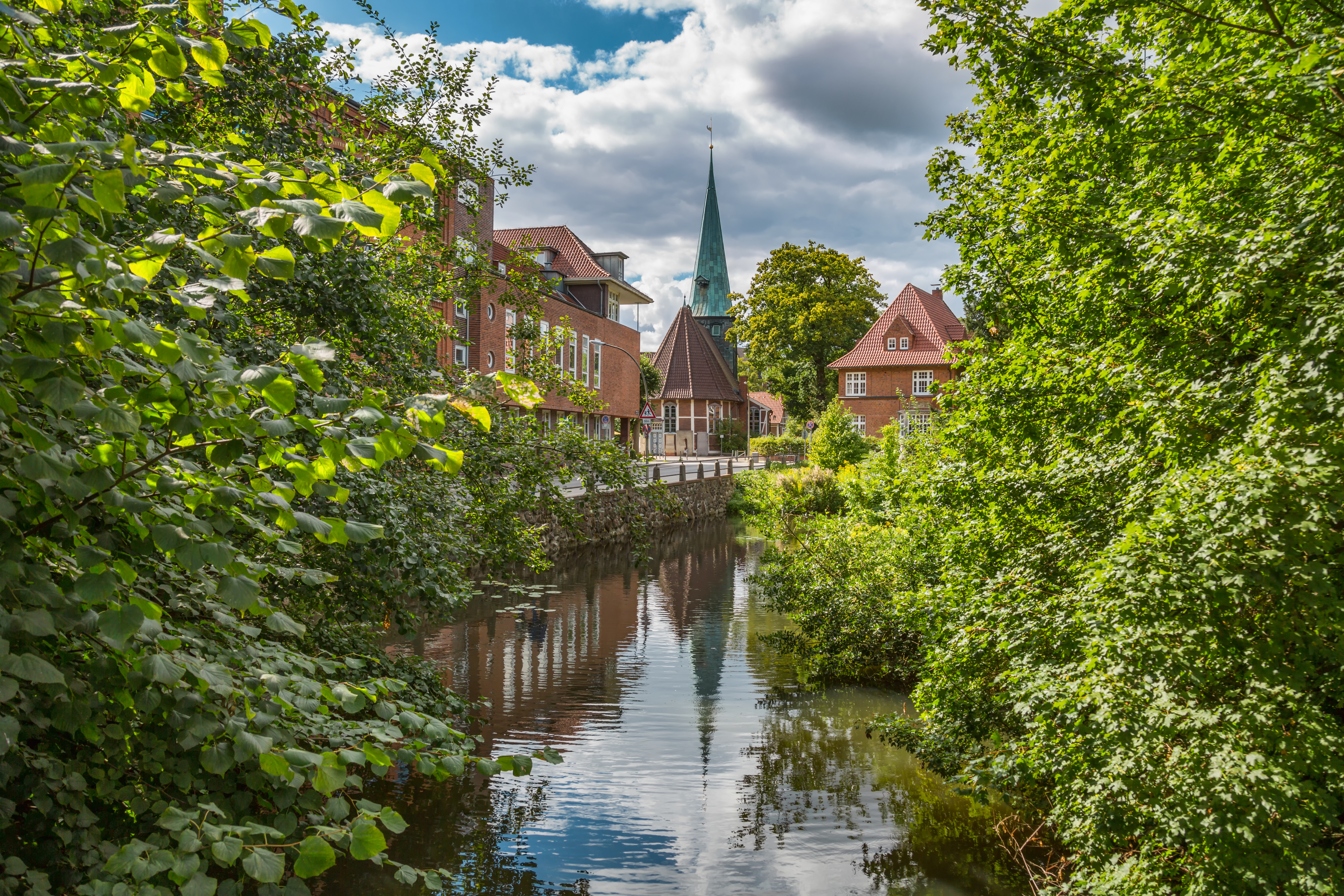 Blick auf einen Fluss mit Wohnhäusern und einer Kirche im Hintergrund, umgeben von Bäumen.