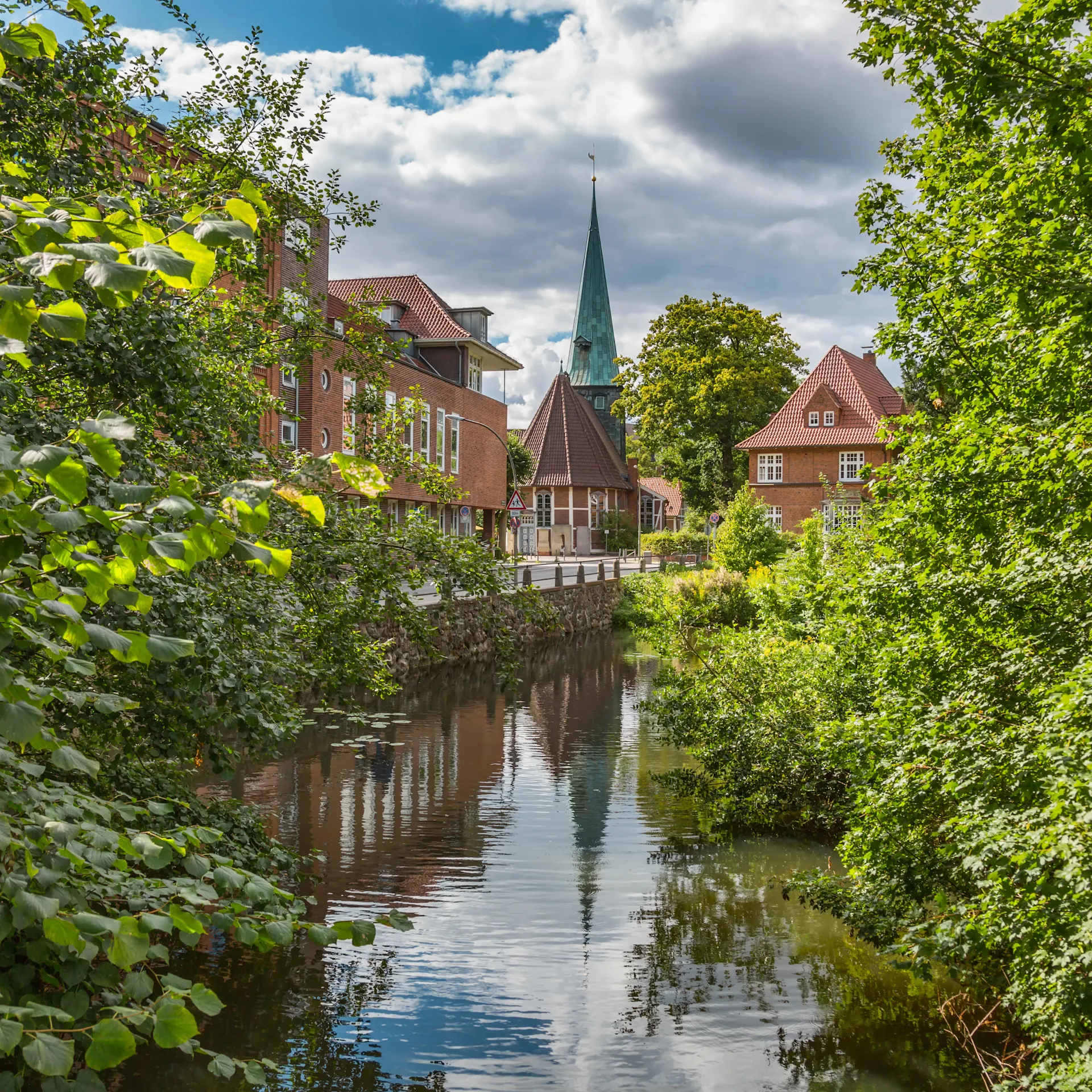 Blick auf einen Fluss mit Wohnhäusern und einer Kirche im Hintergrund, umgeben von Bäumen.
