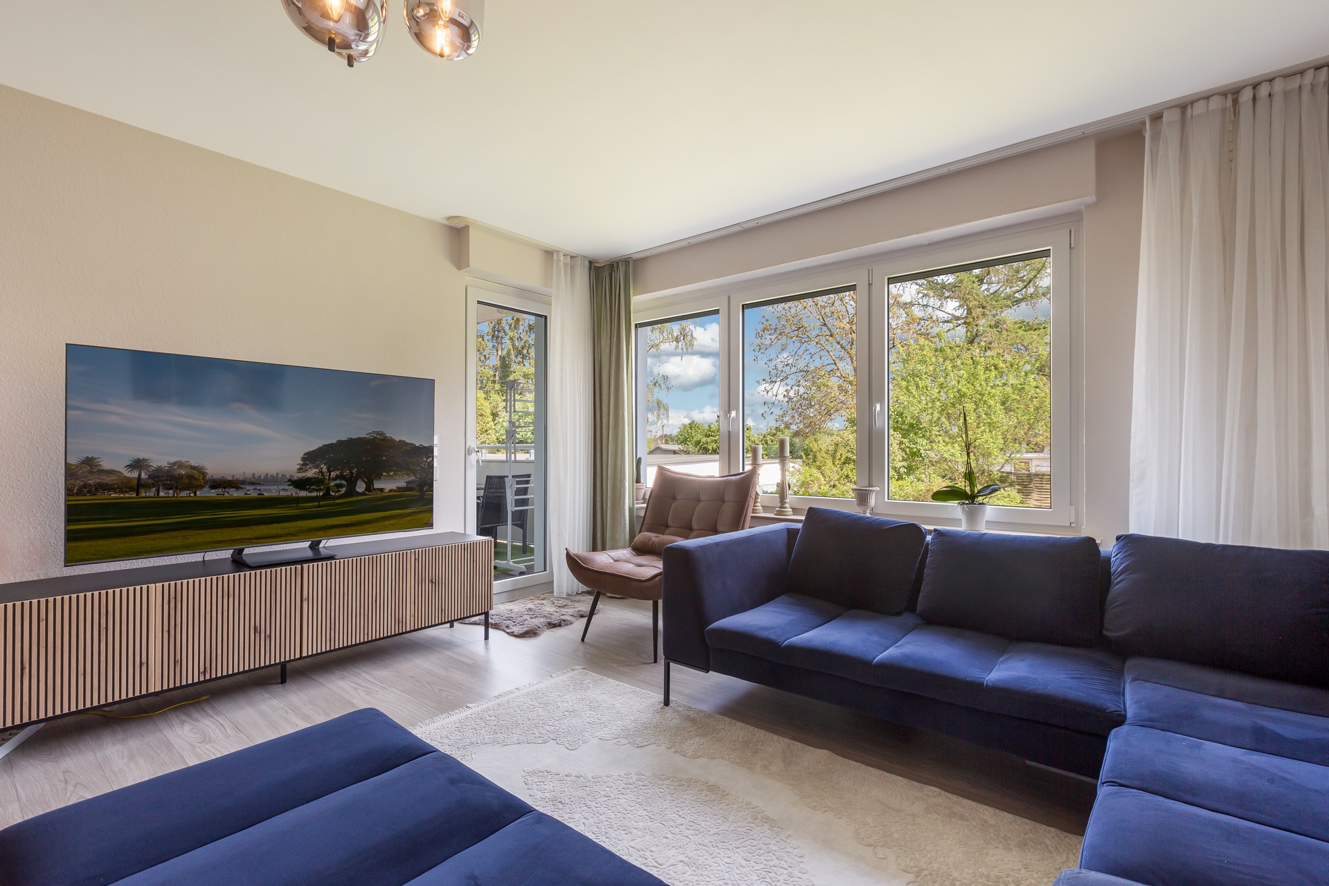 Helles Wohnzimmer mit zwei blauen Sofas, großem Fernseher, Sessel und großen Fenstern mit Blick ins Grüne.