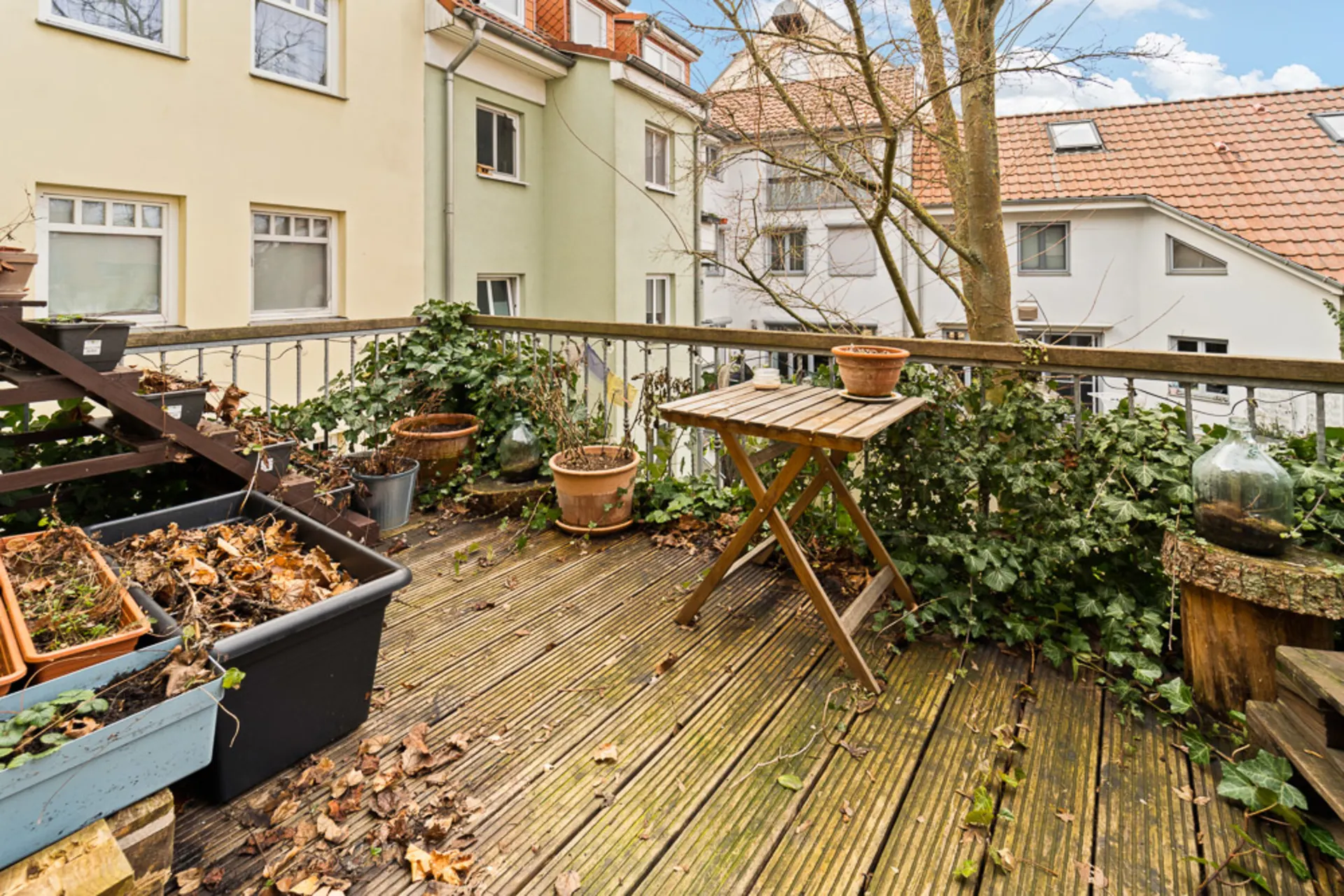 Holzterrasse mit Blumentöpfen, Blättern und einem kleinen Tisch, umgeben von Pflanzen und Wohnhäusern.