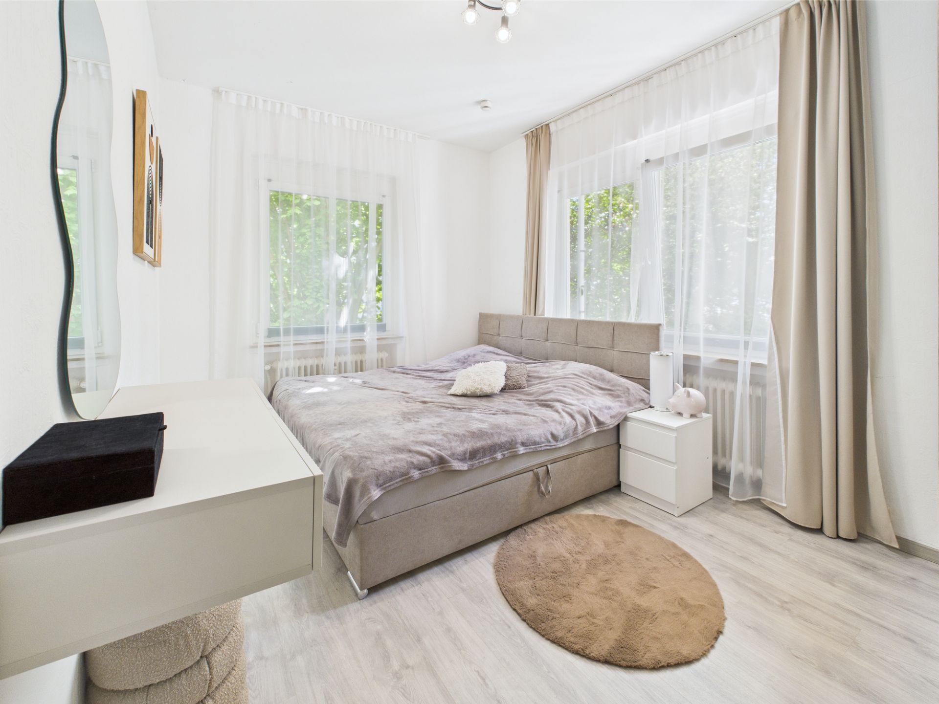 Helles Schlafzimmer mit großem Bett, Nachttisch und zwei Fenstern mit Gardinen.