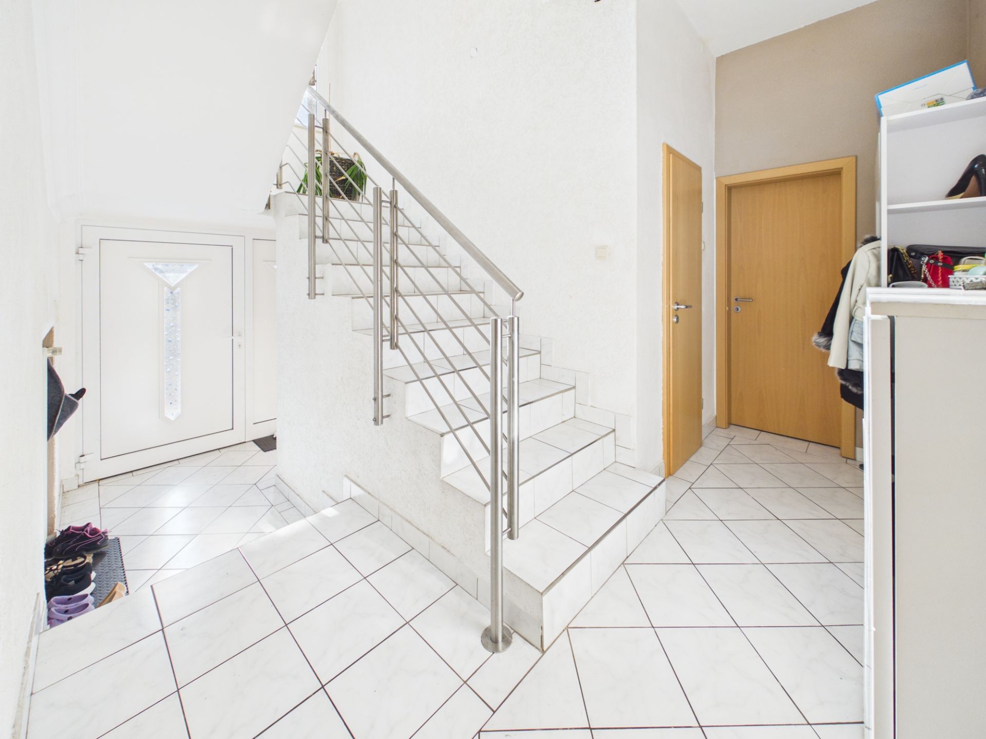 Heller Flur mit weißer Treppe, zwei Holztüren und offenen Regalen.