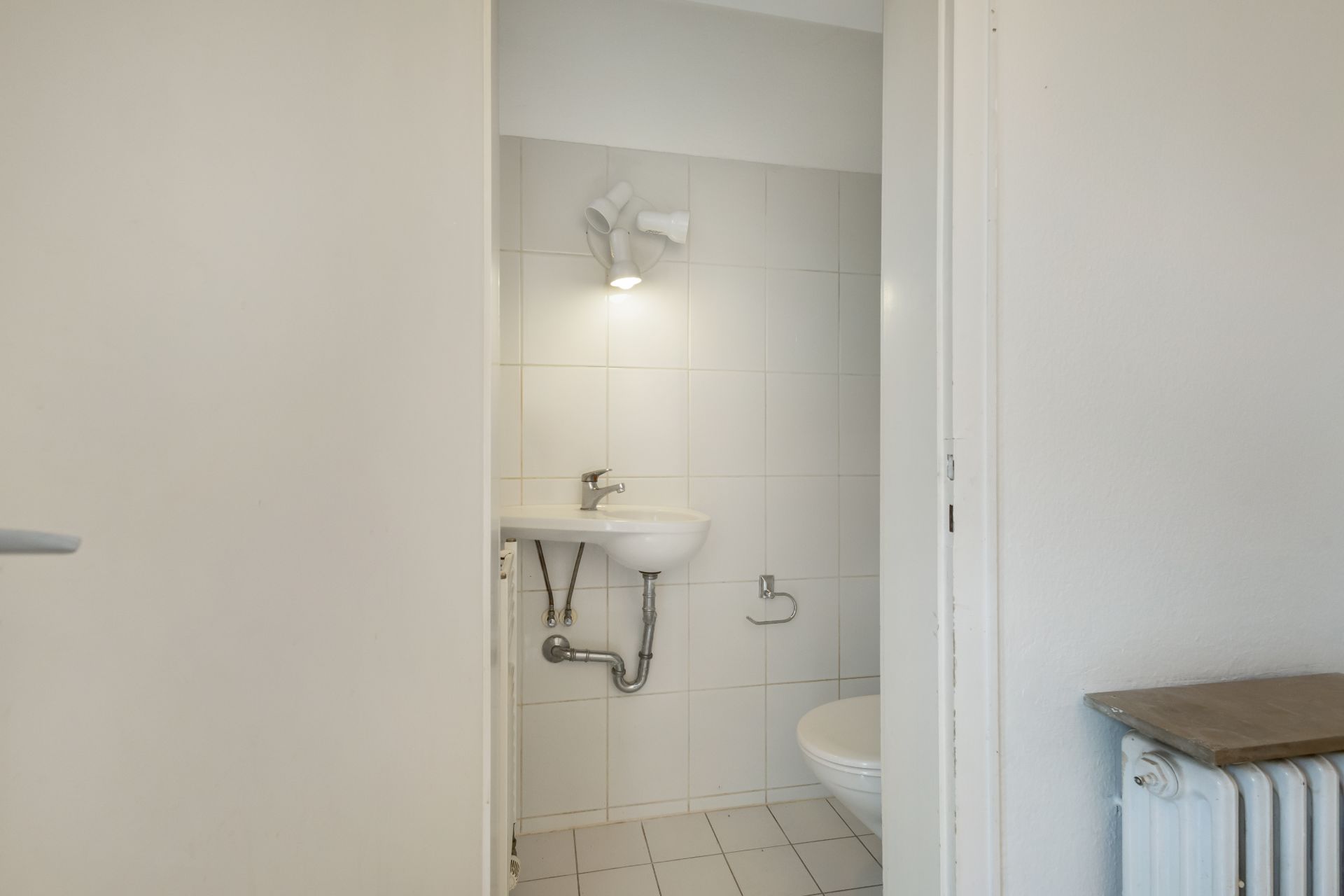 Kleines Badezimmer mit Waschbecken, Toilette und Wandlampe, weiße Fliesen an Wand und Boden.