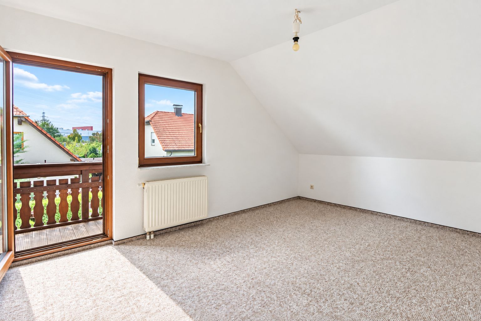 Helles Zimmer mit Teppichboden, Fenster, Tür zum Balkon und schräger Decke.