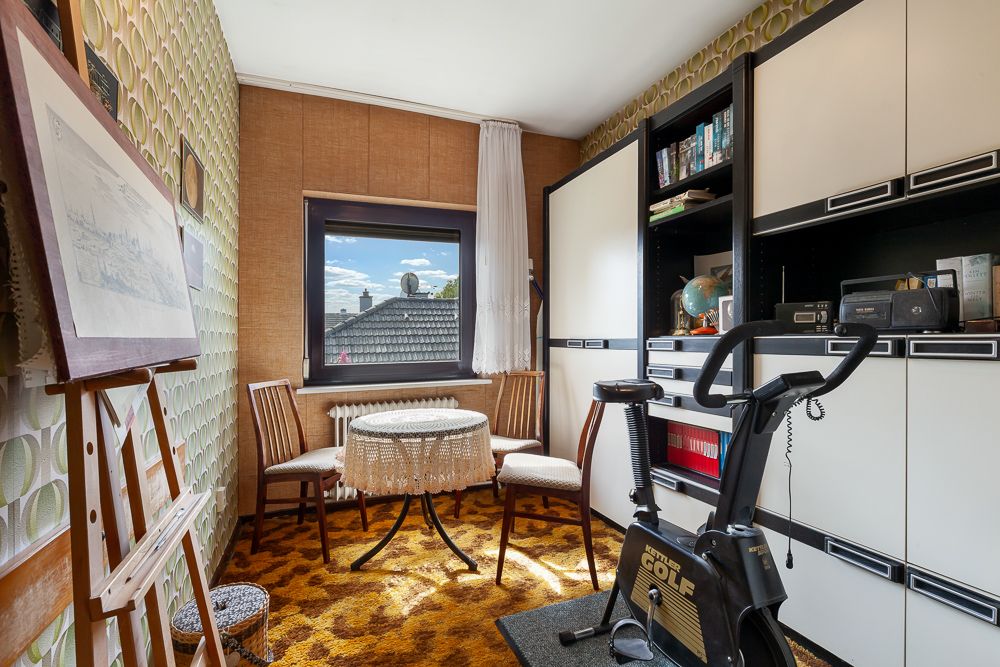 Kleines Zimmer mit Teppich, Tisch, drei Stühlen, Regalwand und Heimtrainer, Fenster mit Blick aufs Dach.