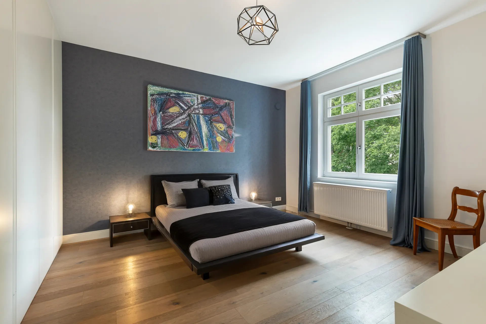 Schlafzimmer mit Doppelbett, Nachttischen, großem Fenster, Vorhängen und modernem Wandbild.