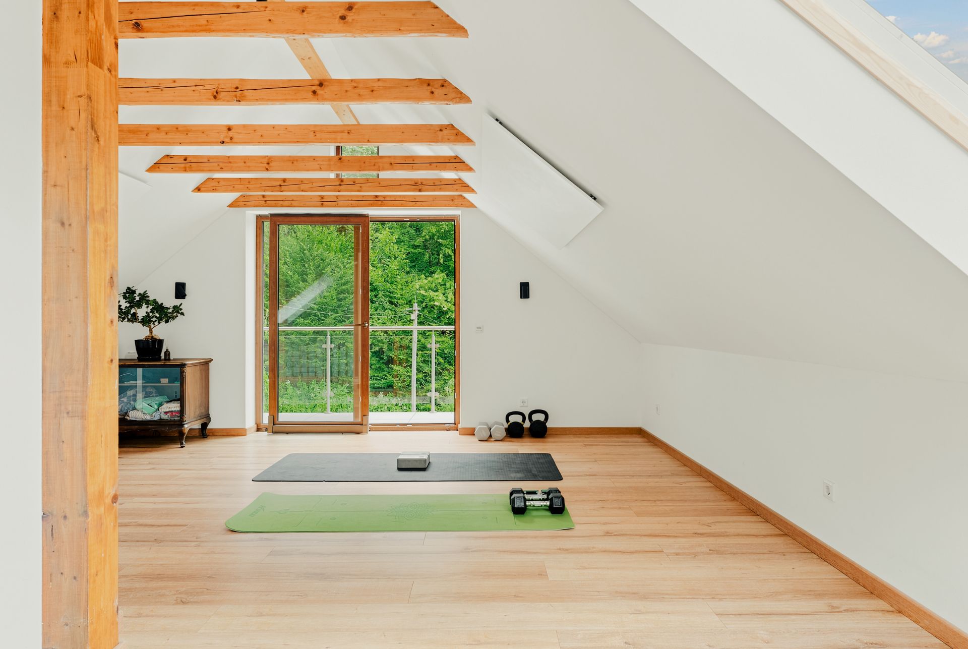 Heller Dachbodenraum mit Holzbalken, Sportmatten, Hanteln und Blick auf einen Balkon mit viel Grün.