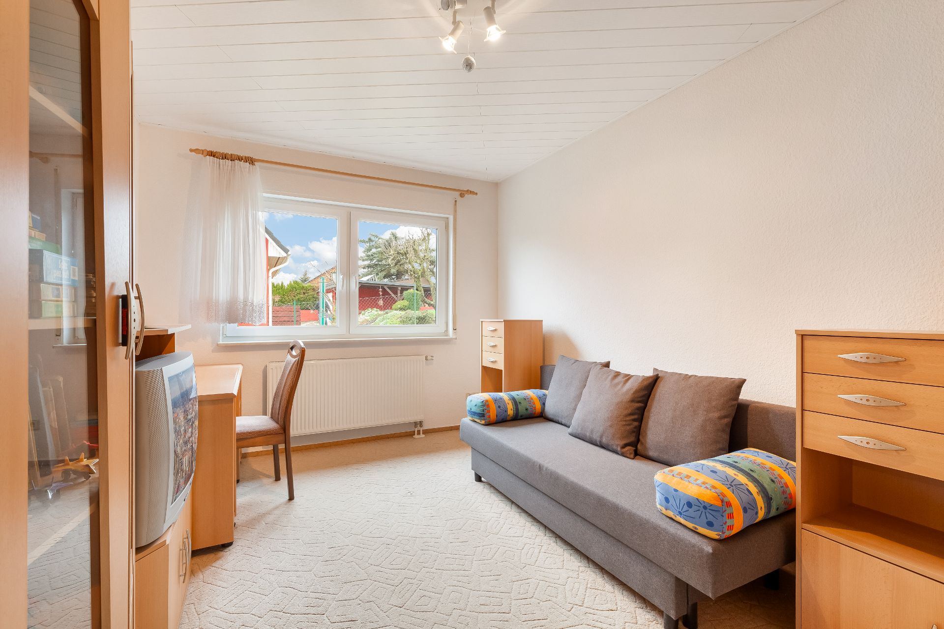 Helles Zimmer mit grauem Sofa, Holzschränken, Schreibtisch und Fenster mit Ausblick.