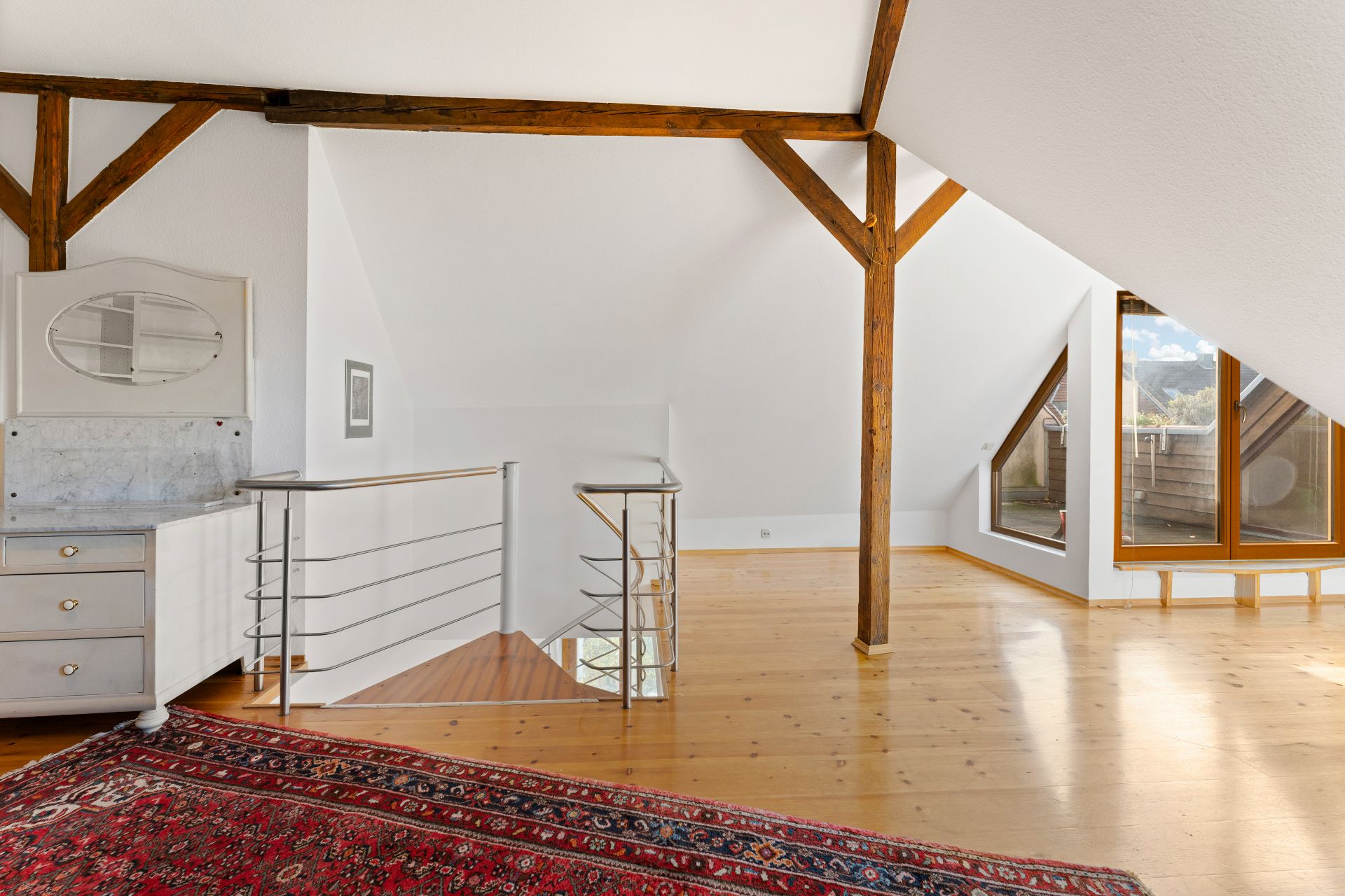 Helles Dachgeschoss mit Holzbalken, Holzfußboden, Fensterfront und rotem Teppich.