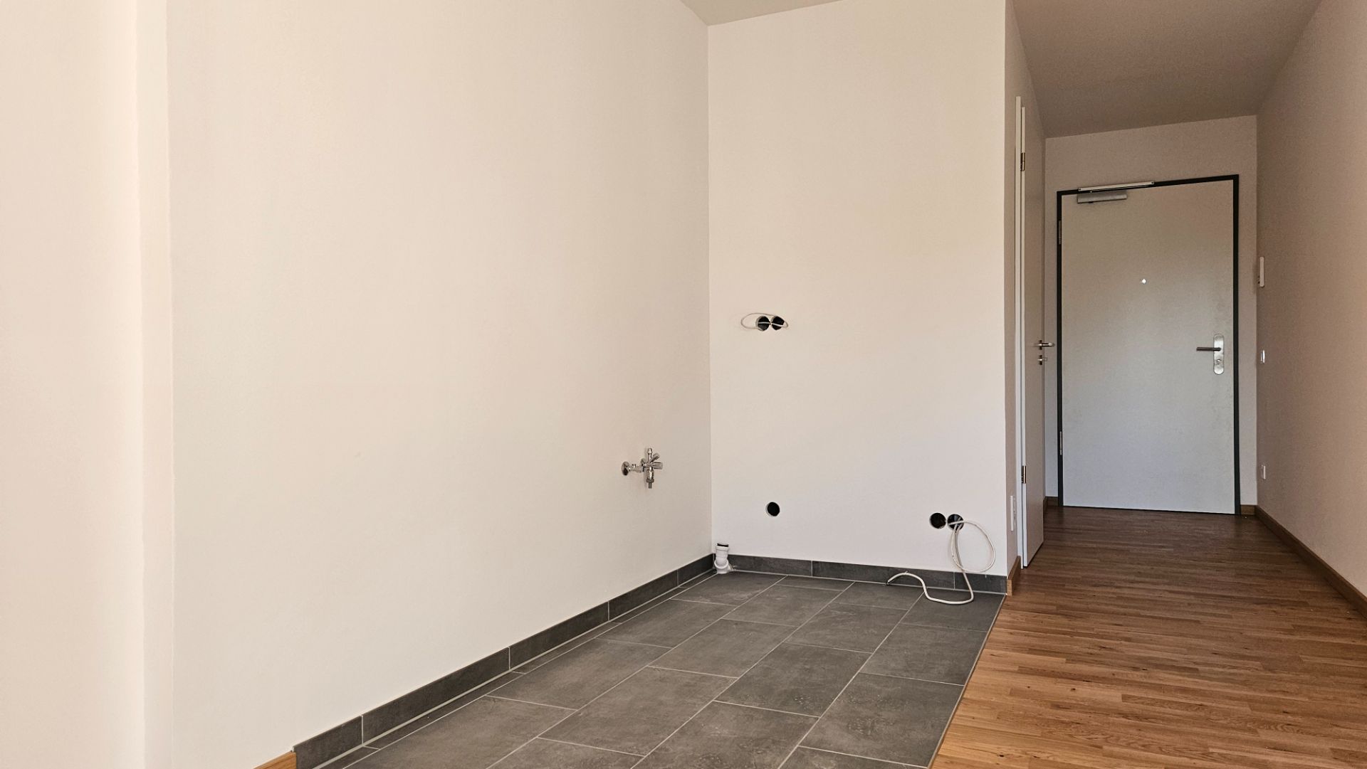 Leerer Raum mit Fliesenboden, Anschlüssen an der Wand und Eingangsbereich mit Holzboden sichtbar.
