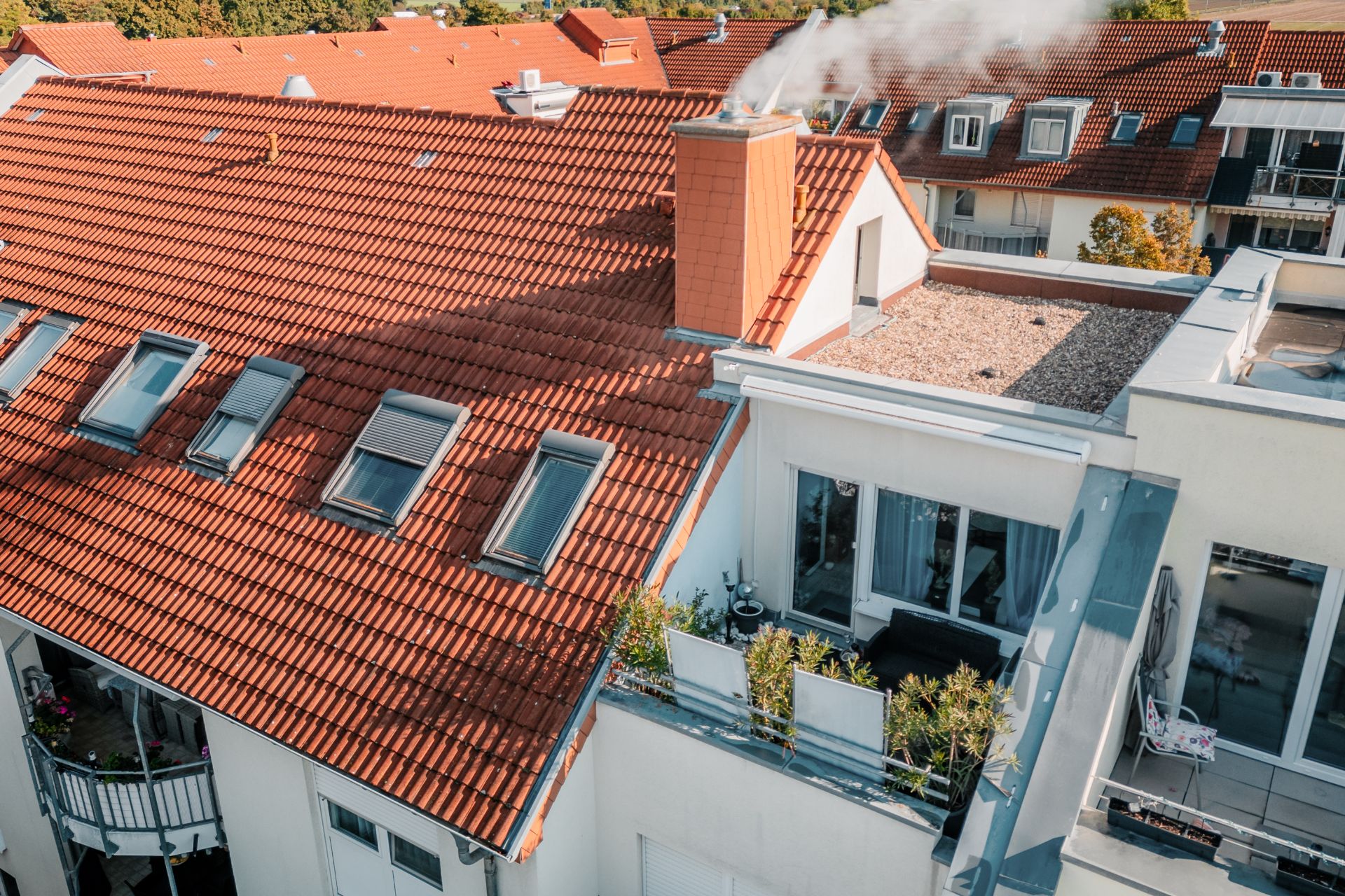 Wohngebäude mit rotem Ziegeldach, Balkon und rauchendem Schornstein.