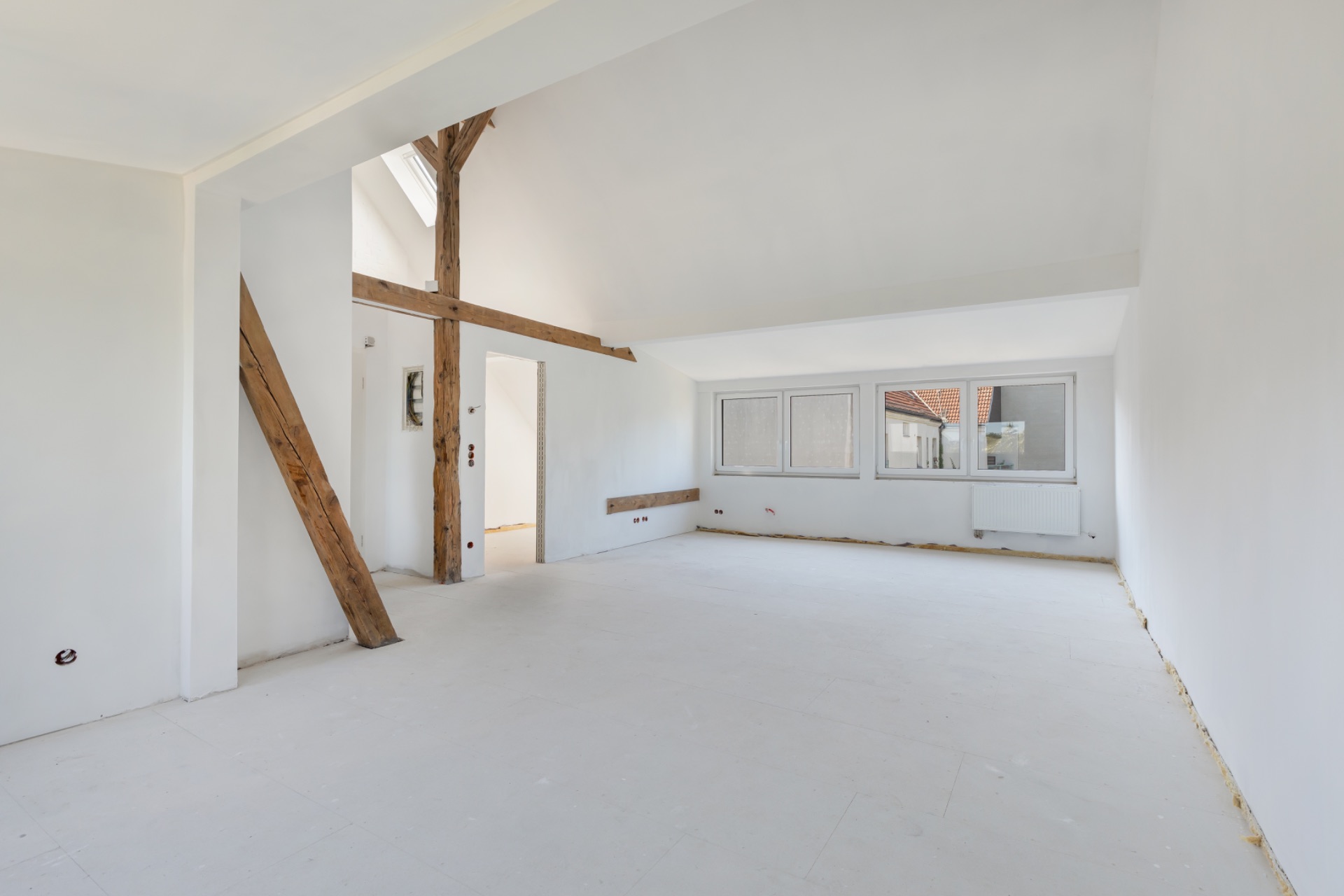Heller, leerer Raum mit weißen Wänden, großen Fenstern und sichtbaren Holzträgern.